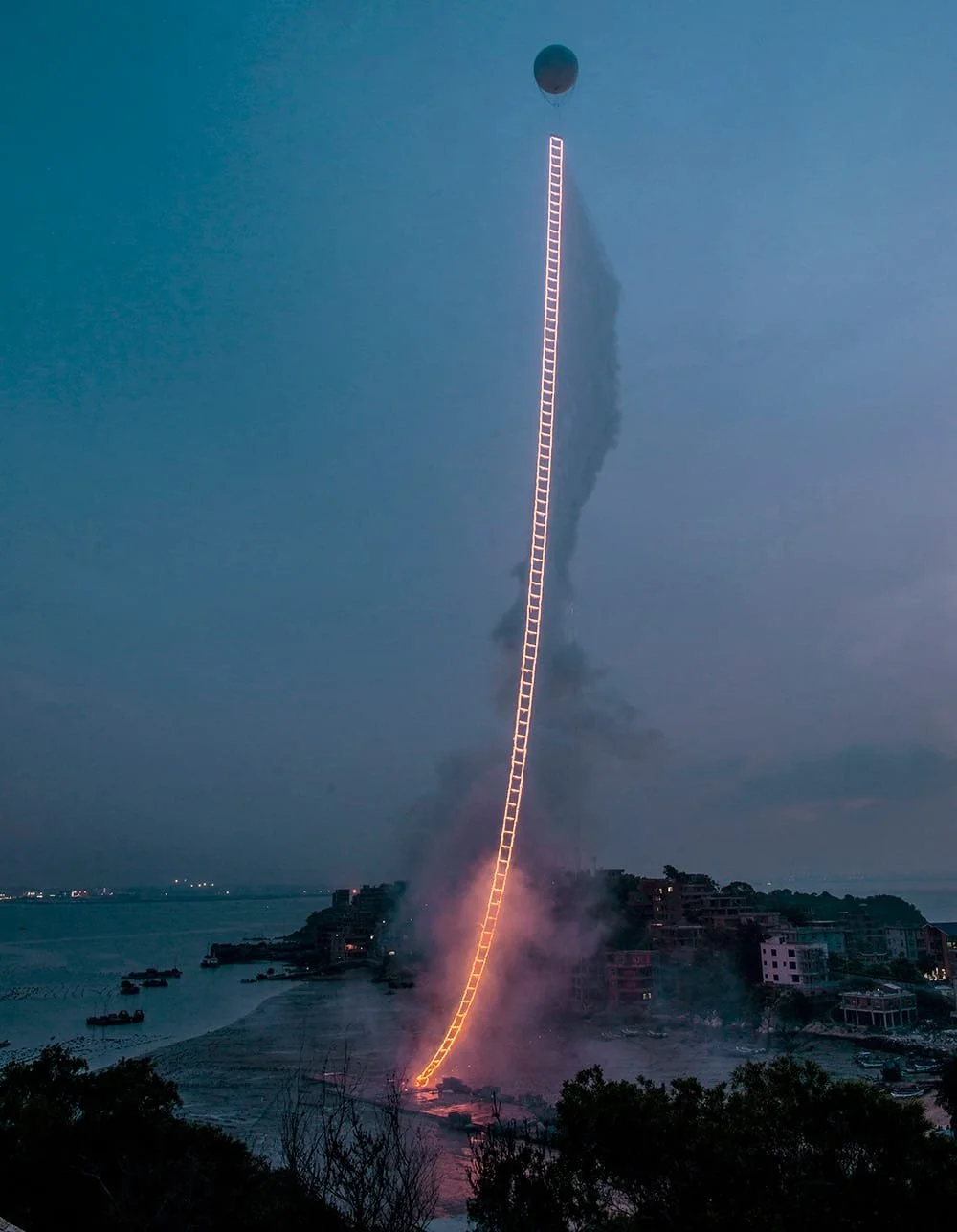 A rocket is launching vertically with a trail of bright orange flames and smoke, reaching into the dusk or dawn sky, near a body of water with boats and buildings visible in the background.