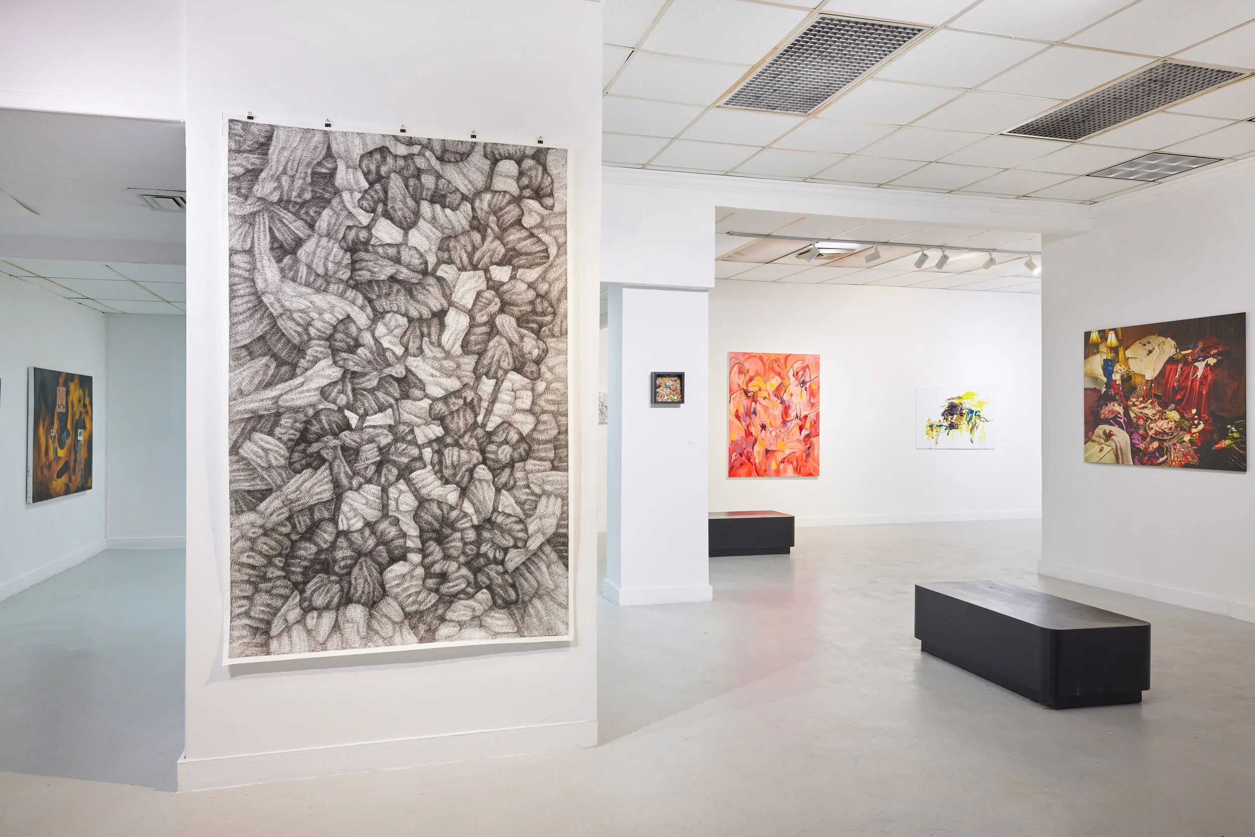 An art gallery with white walls and a white drop ceiling, displaying various paintings. A large, detailed black-and-white artwork of numerous shoes is in the foreground. Smaller colorful paintings are on the walls in the background, along with black benches on the light-colored floor.