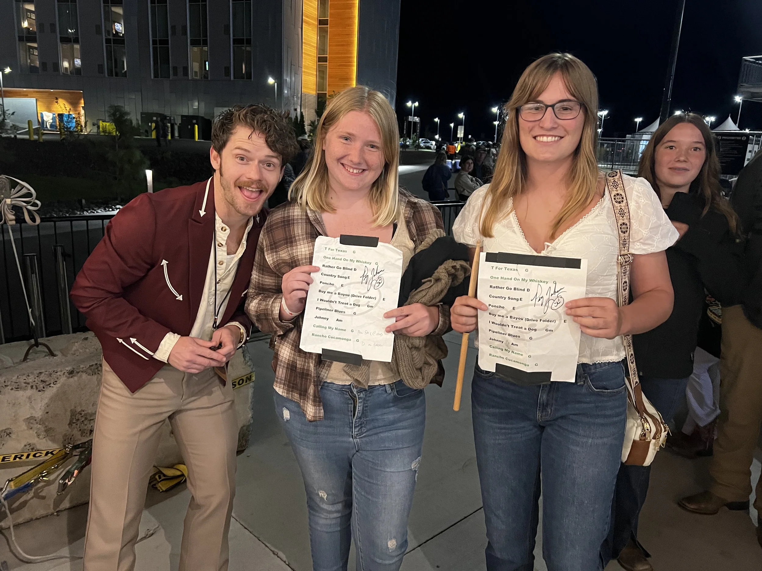 Four people standing outdoors at night, two women holding papers with a list. The group is smiling and posing for the photograph. There are tents and parked cars in the background.