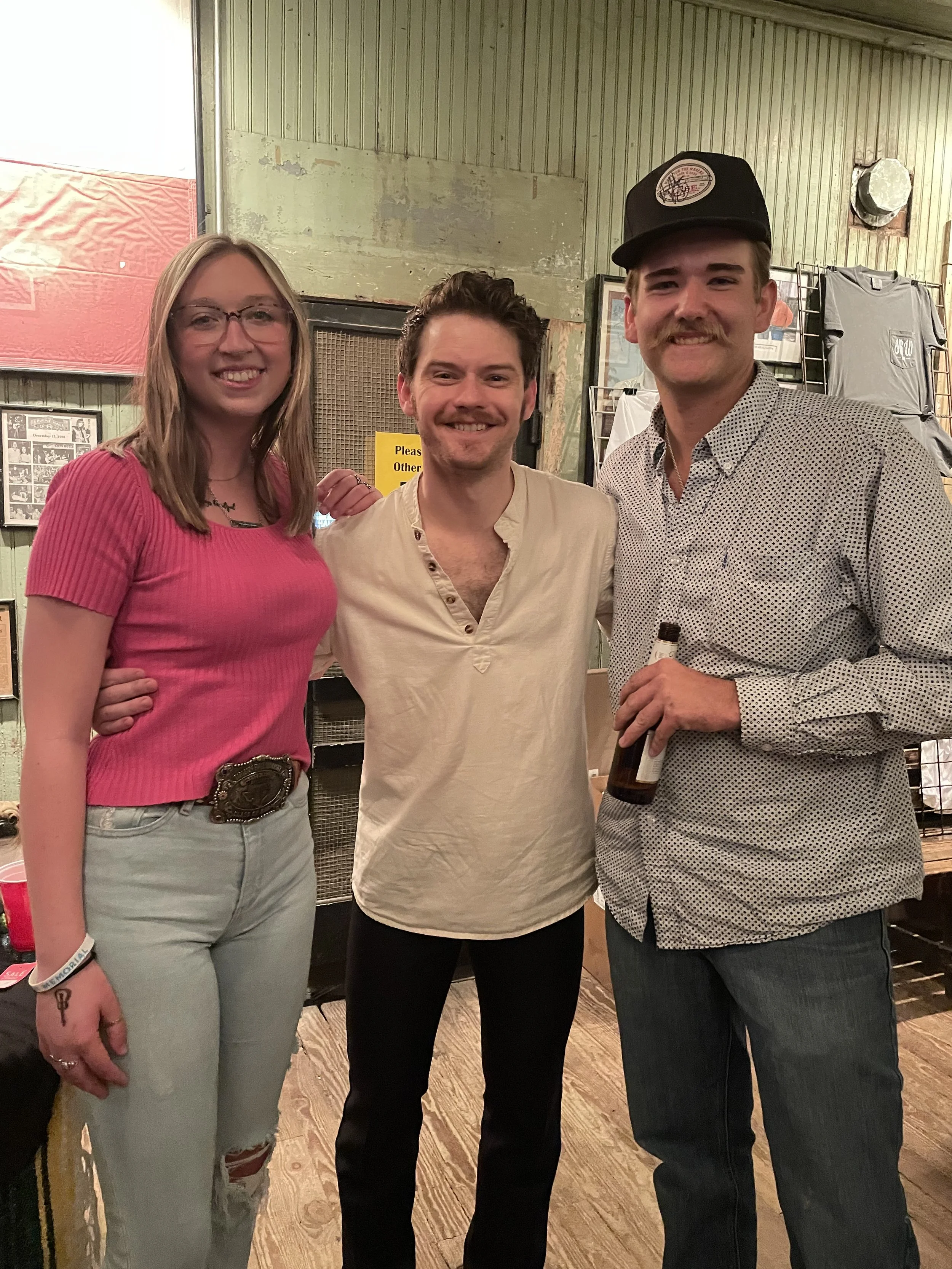 Three people smiling and posing for a photo indoors. The person on the left is a woman with glasses and blonde hair, wearing a pink top and light-colored jeans. The person in the middle is a man with short dark hair, wearing a beige shirt. The person on the right is a man with a mustache, wearing a black cap and a patterned button-up shirt, holding a bottle in his right hand.