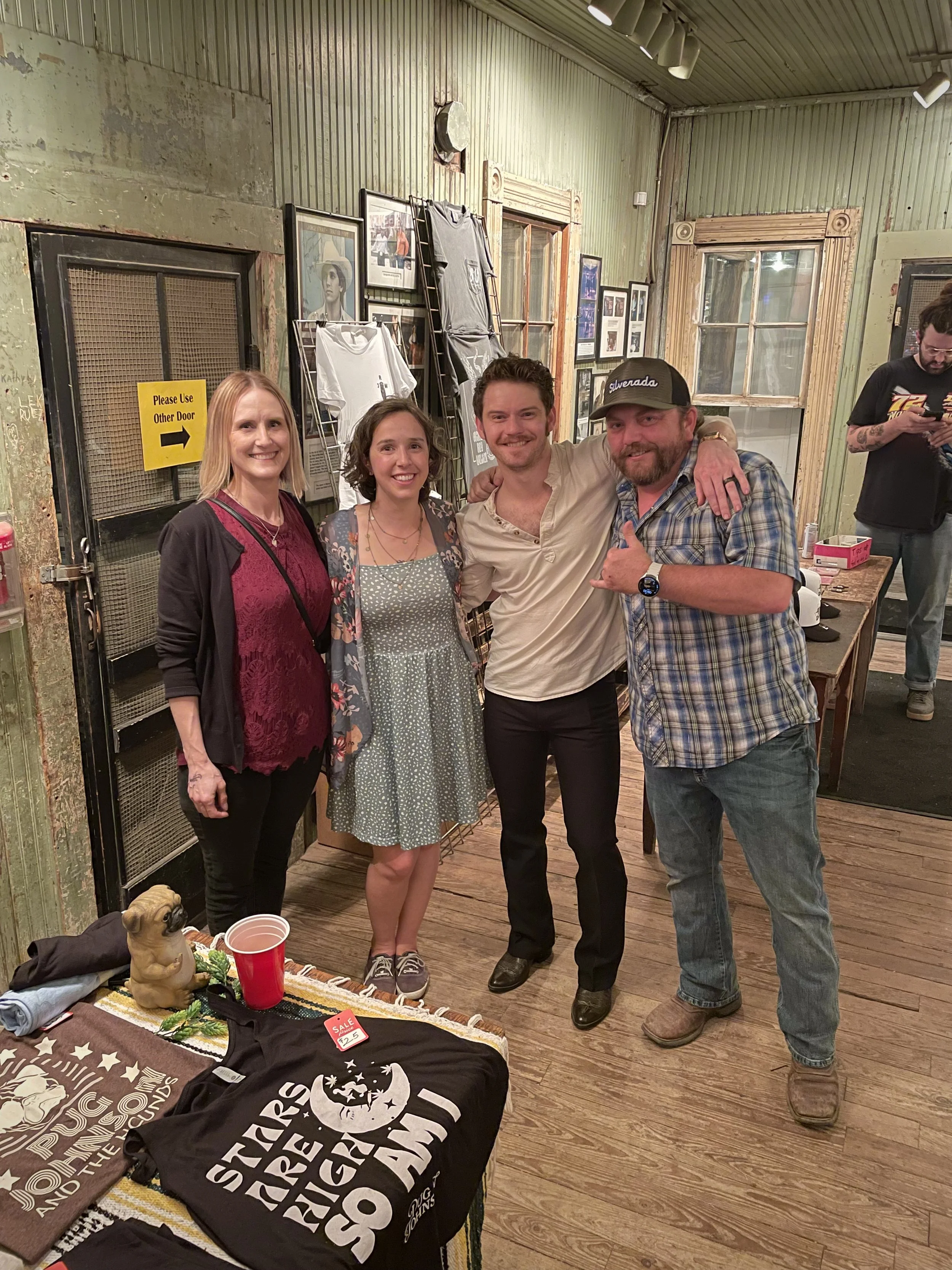Four people standing together in a room, smiling at the camera. The room has green walls, wooden trim, and framed pictures hanging. A table with various merchandise, including T-shirts and a decorative dog figure, is in the foreground.