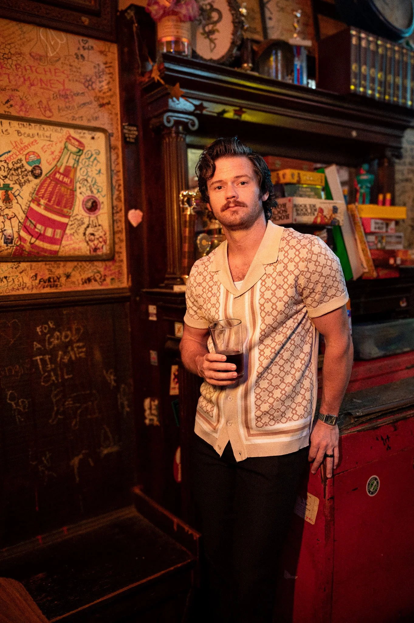 A man with dark hair and a mustache wearing a patterned short sleeve shirt, holding a glass of dark beverage, standing in a dimly lit room with colorful artwork and books.