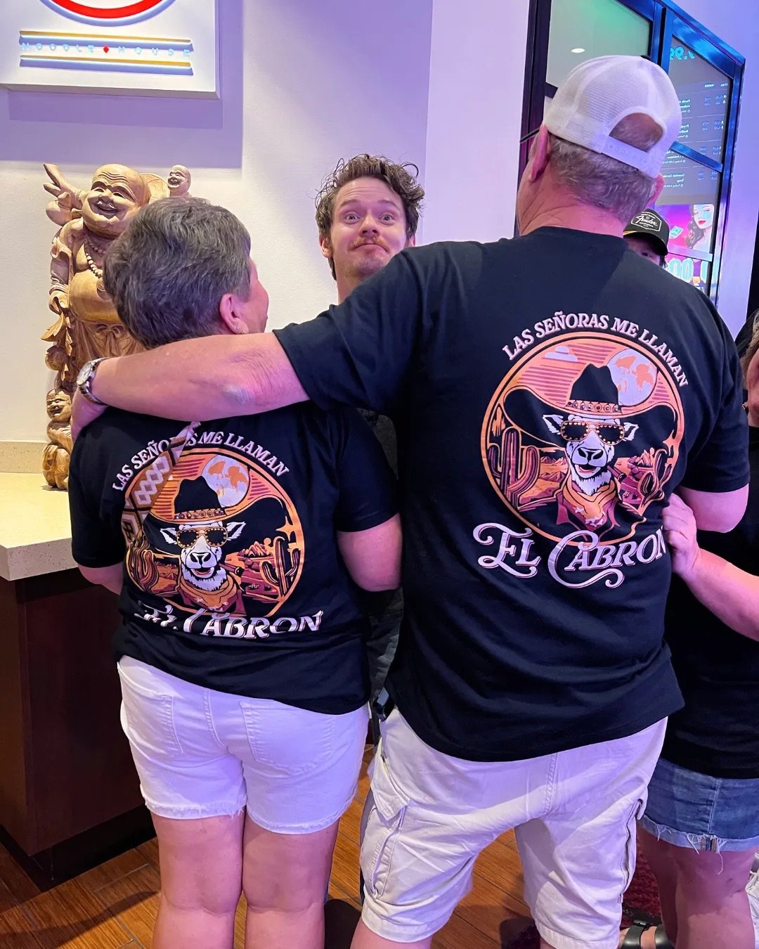 Three people are standing indoors, wearing matching black T-shirts with a llama illustration on the back. The llama is wearing sunglasses and a hat, with the text "Las Señoritas Me Llaman El Cabron". One person has their arm around another, and the person in the middle is making a funny face. There is a wooden sculpture of a laughing Buddha and purple-lit screens in the background.