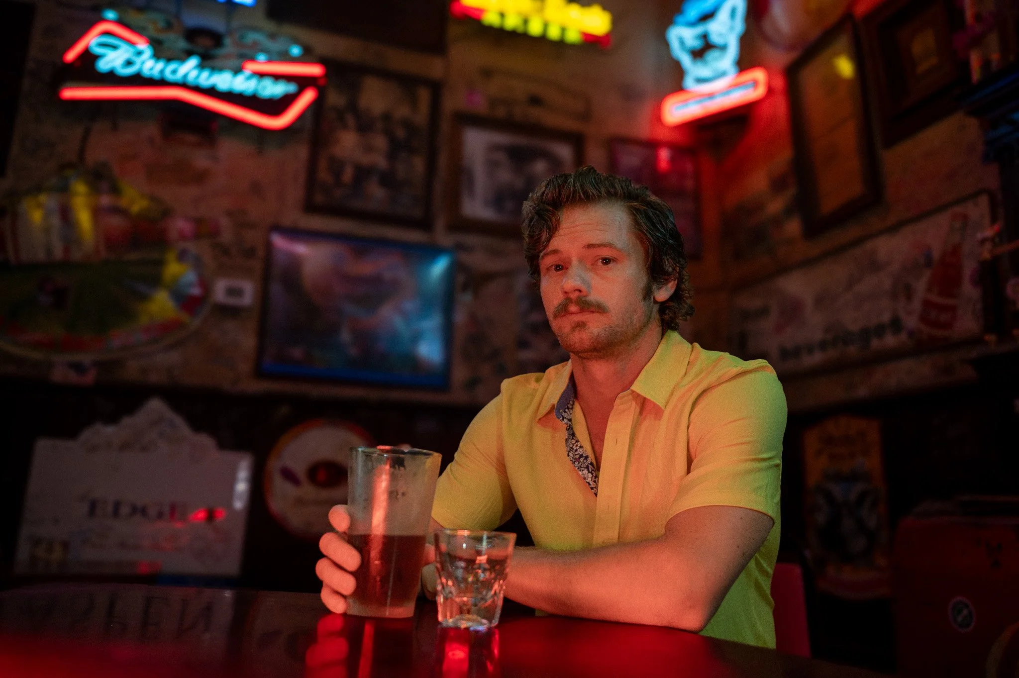 A man with a mustache and curly hair wearing a yellow shirt sitting at a table in a dimly lit bar or restaurant, holding a glass of dark beer. The background has graffiti and illuminated neon signs.