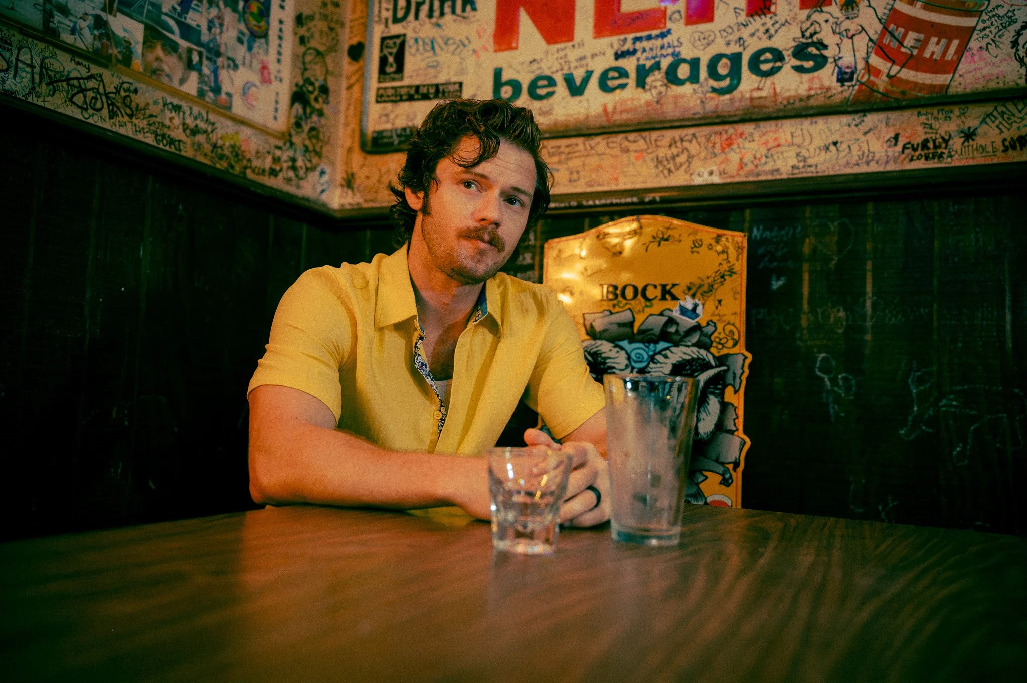 A man with dark curly hair and a mustache sits at a bar or restaurant table, wearing a yellow shirt with a floral collar. There are multiple empty glasses in front of him on the table, and he is looking off to the side. The background features wall art covered in numerous graffiti and posters, including a large sign that reads 'drinks' and a poster with the word 'beverages'.