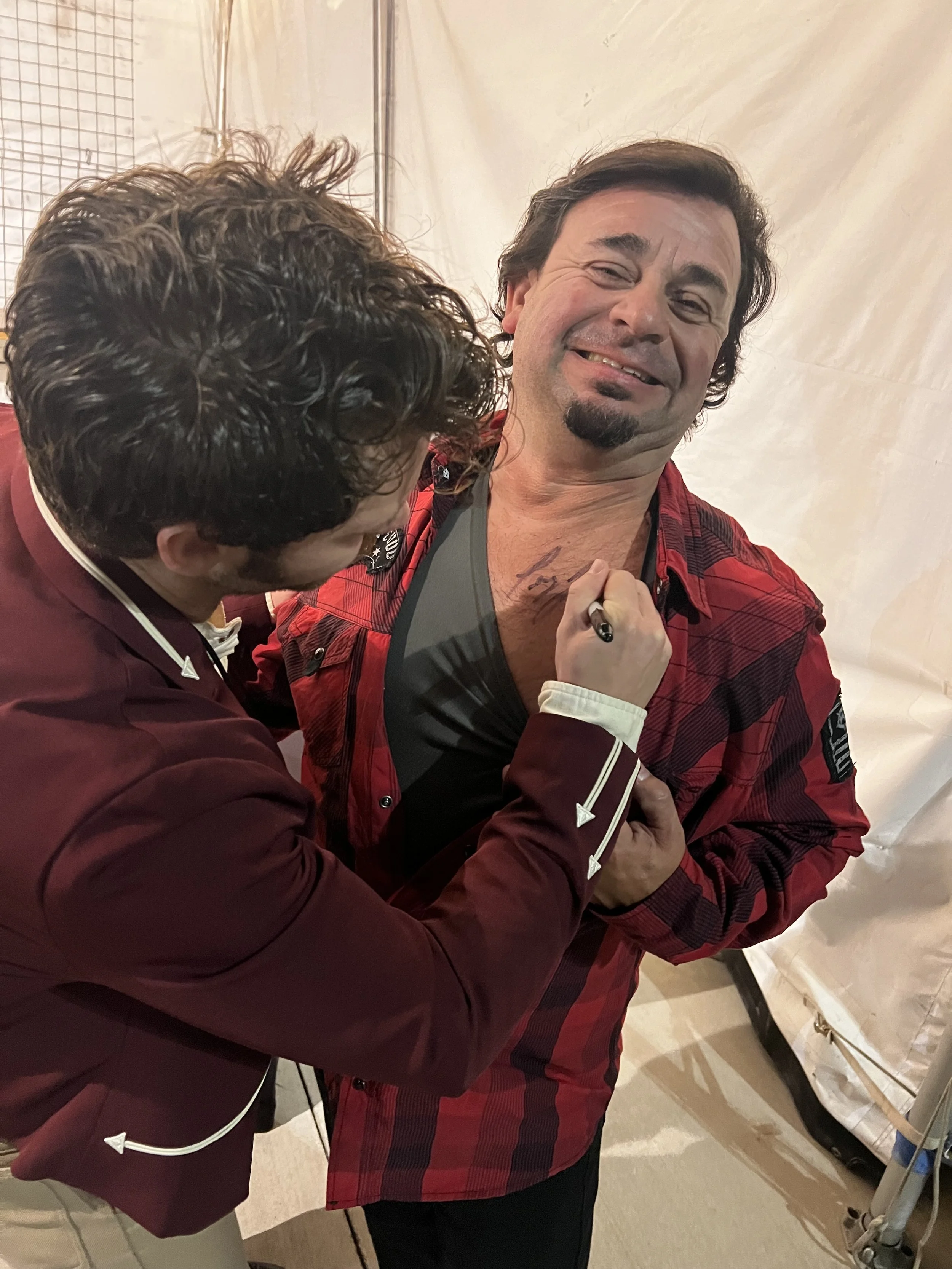 A man with dark, curly hair and a goatee wearing a red plaid jacket and black shirt, giving a mustache to another person with curly hair, in a close-up indoor setting.