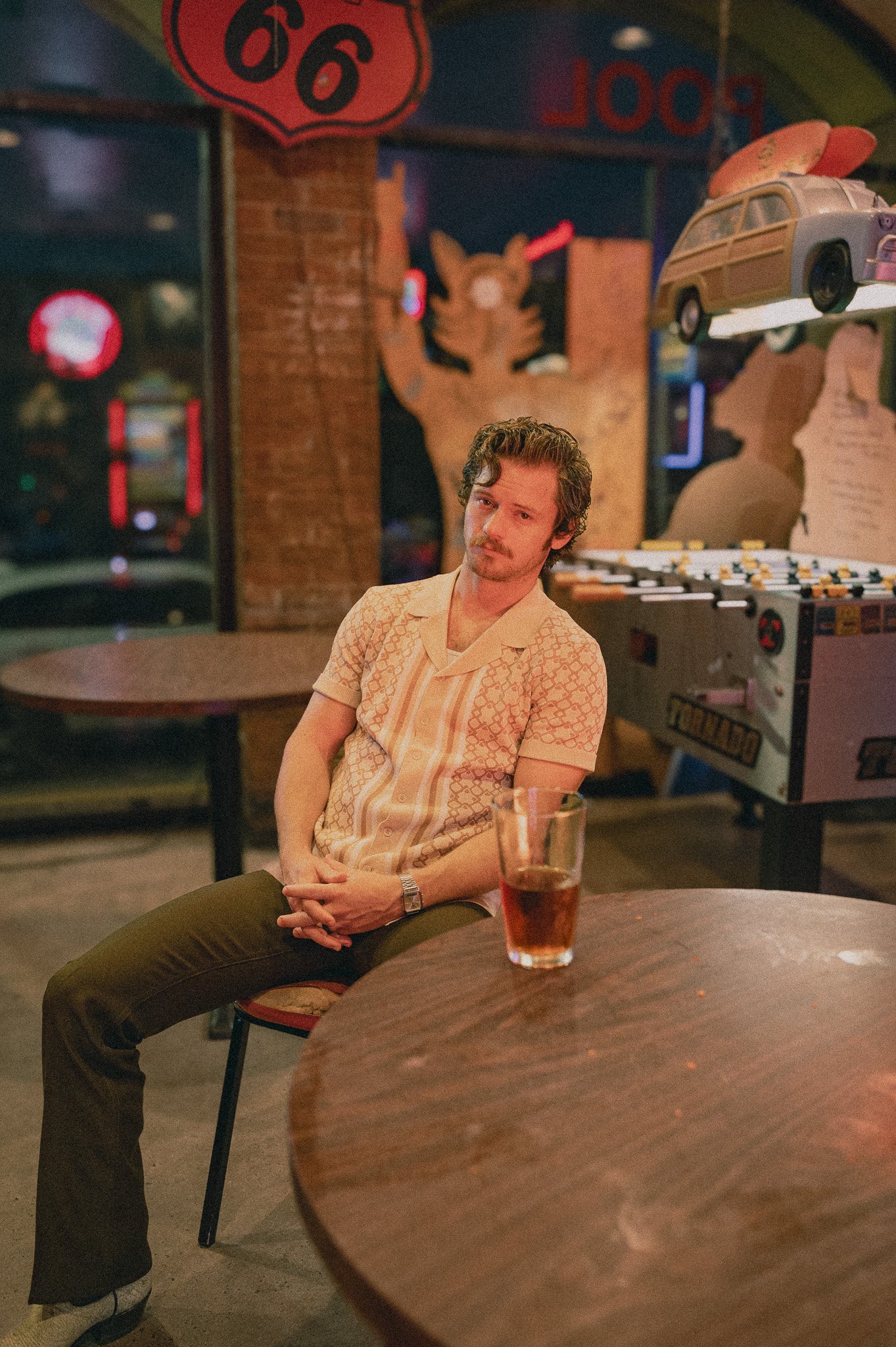 A man with dark curly hair, a mustache, and a beard, sitting at a round wooden table in a bar or restaurant. He is wearing a short-sleeved, patterned shirt in shades of yellow and orange, and dark pants. In front of him on the table is a glass of dark soda with ice. The background has brick walls, a vintage car model hanging from the ceiling, a fox-shaped wall decoration, and a neon sign.