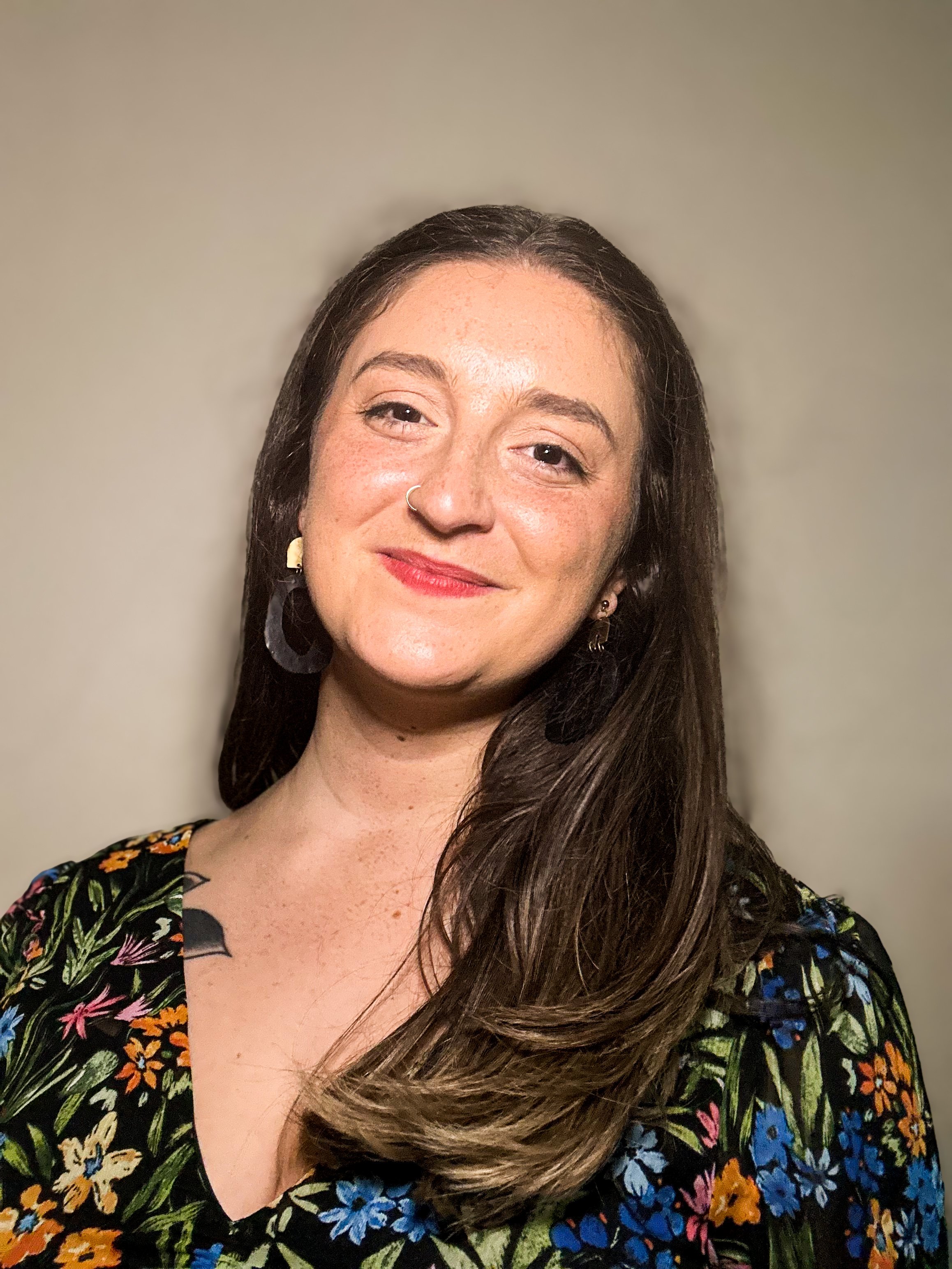 A woman with long brown hair, wearing a floral top, smiling and posing against a plain beige background.