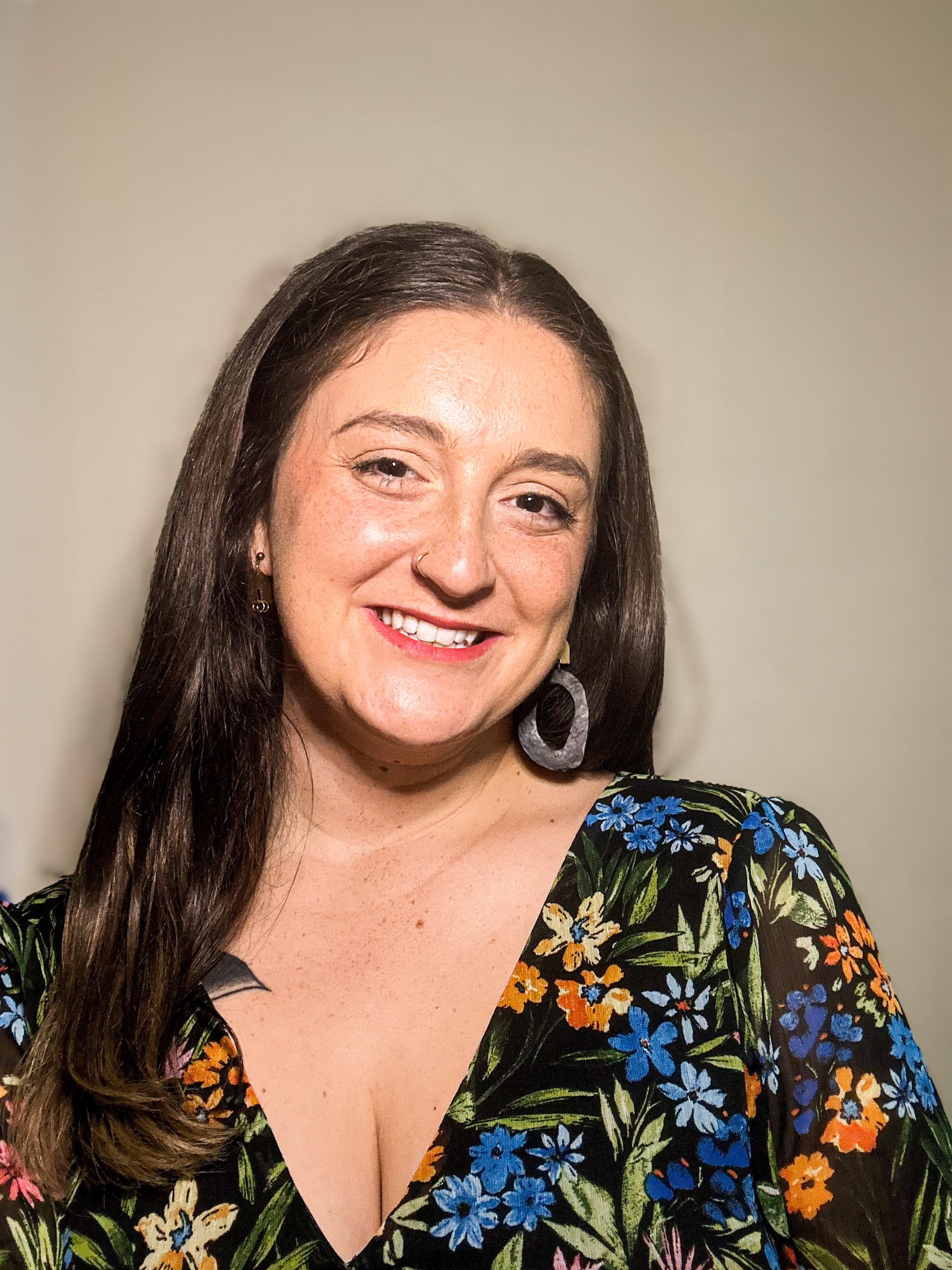 Close-up of a woman with long brown hair, wearing a floral dress with blue, orange, and green colors, smiling and showing her teeth, against a plain beige background.