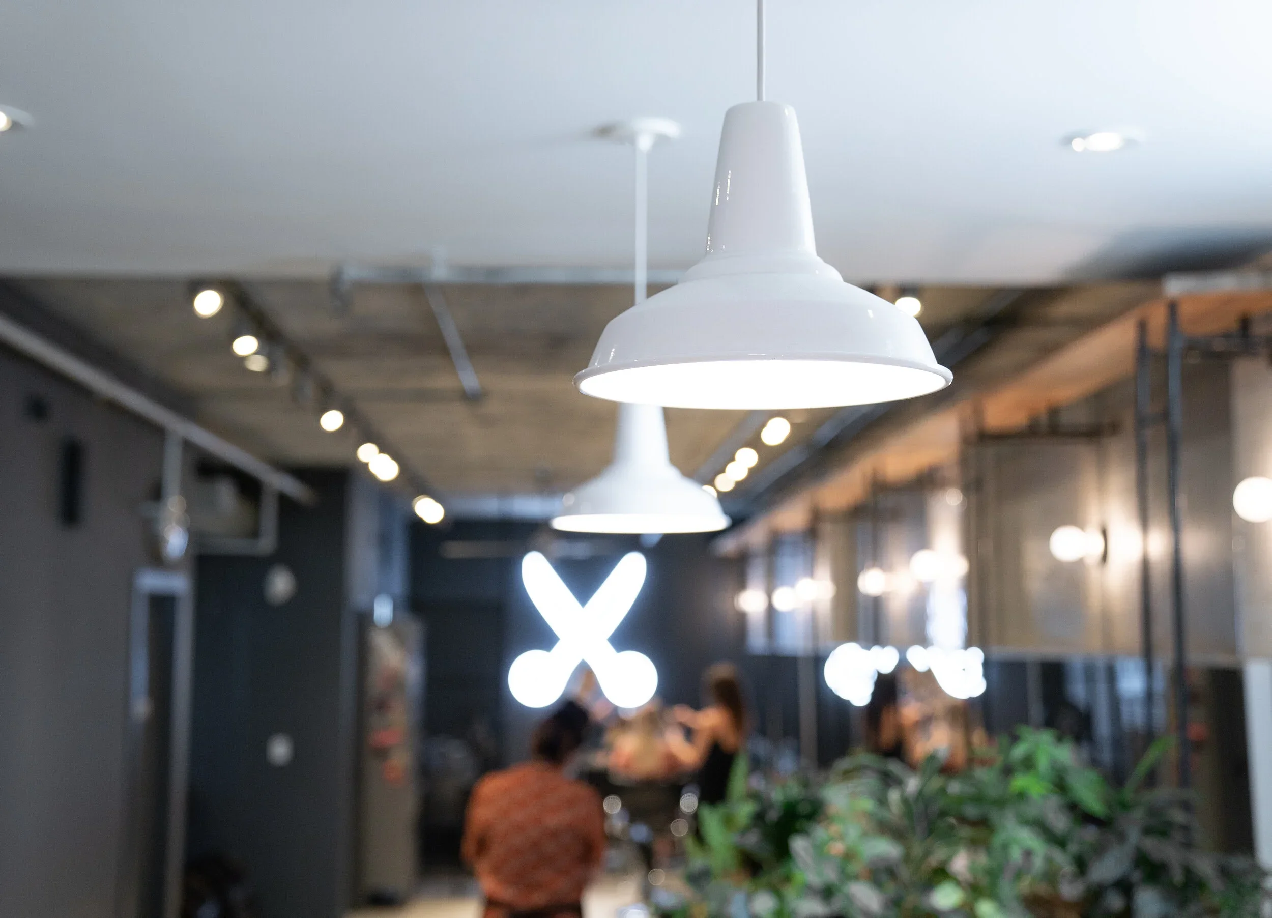 Interior view of Weekly Hair with white hanging pendant lights, some greenery in the foreground, and people dining at tables in the background.