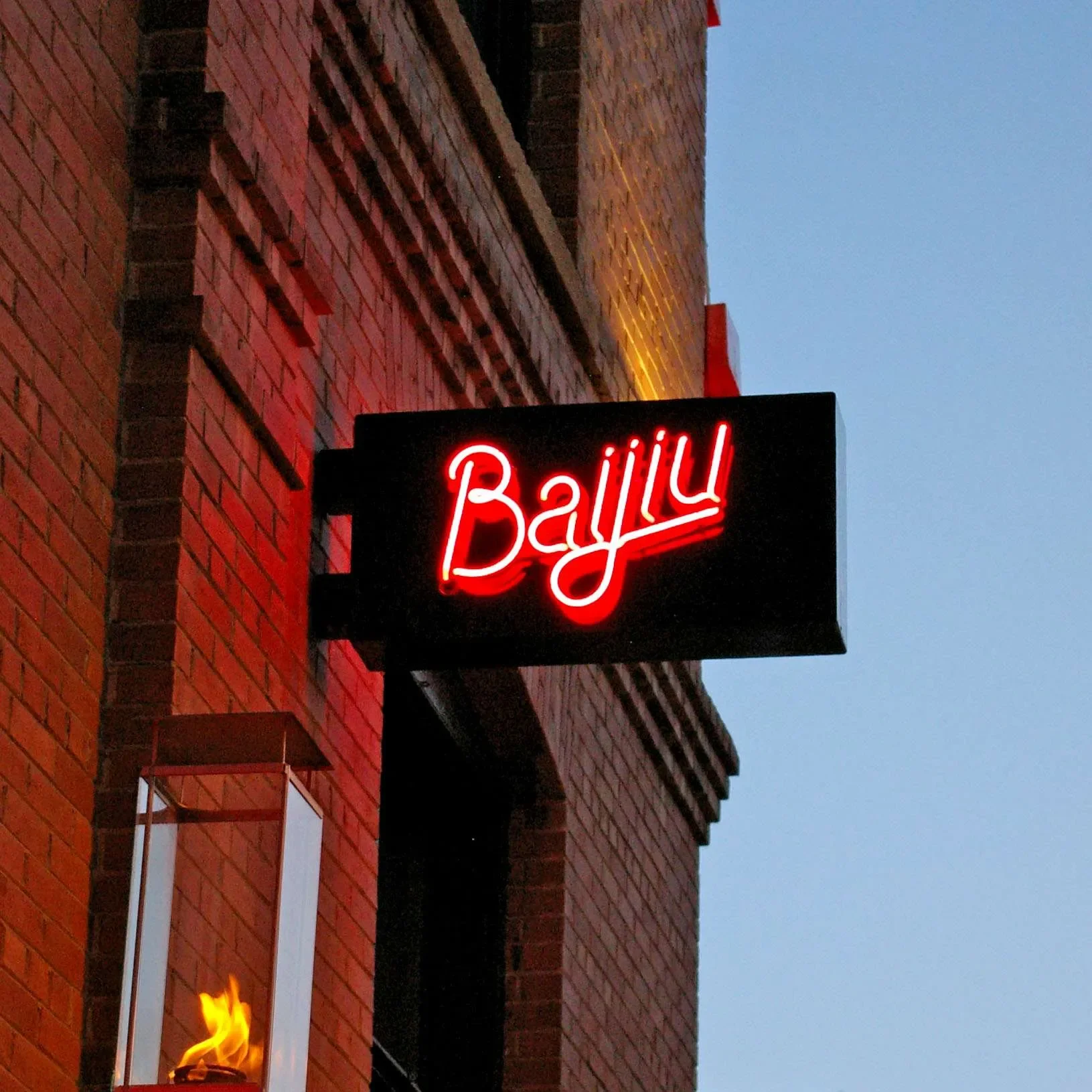 Neon sign reading "Baijiu" mounted on the side of a red brick building, with a fire in a lantern beneath it, against a twilight sky.