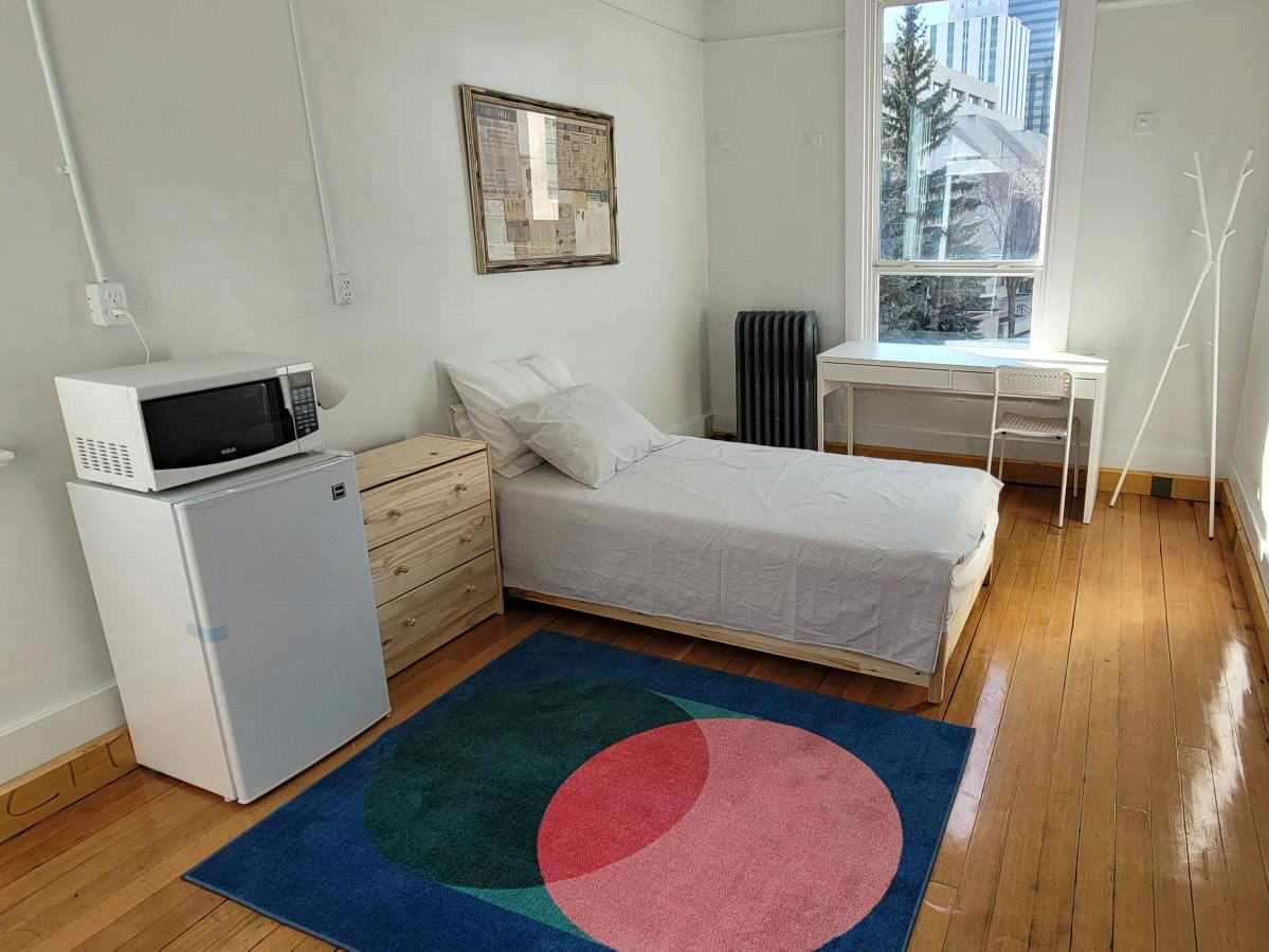 A small, minimalist bedroom with a single bed, white bedding, and a pillThere is a small wooden nightstand, a mini refrigerator with a microwave on top, a colorful geometric rug, a large window with a view of trees and buildings, and a white desk with a chair. A black radiator and a white coat rack are also visible.