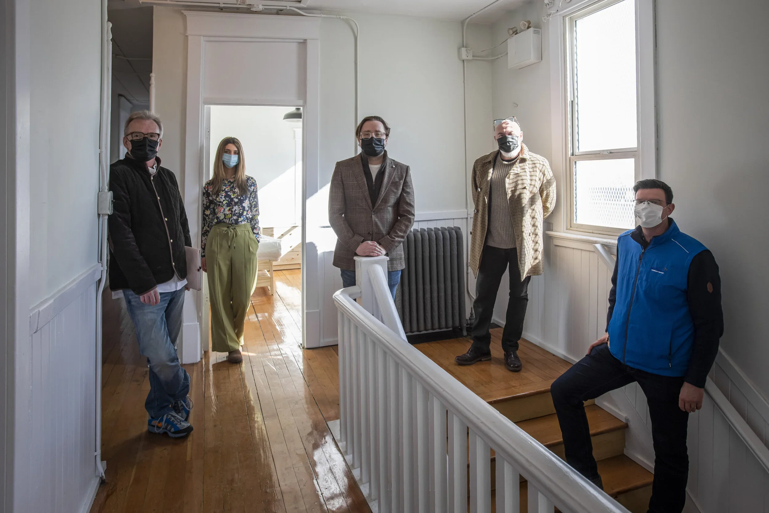 Five people wearing masks standing on a staircase landing in a house, with a woman standing in the open doorway behind them.
