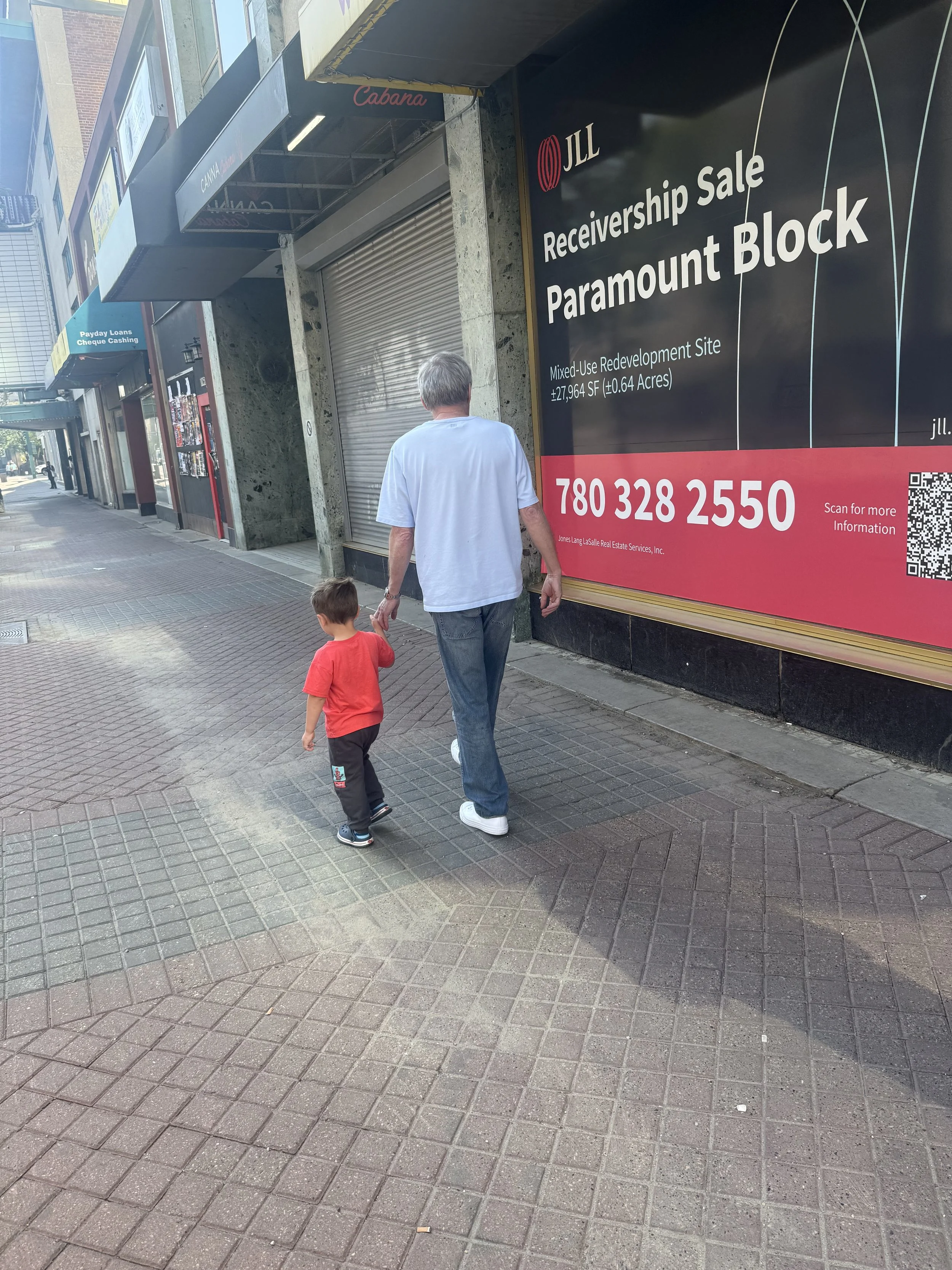 A man and a young boy walking on a sidewalk in an urban area with store signs and a large billboard advertising a property development site.