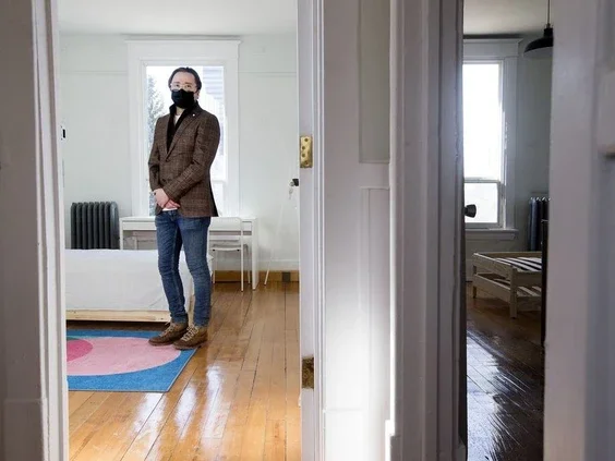 A person wearing a brown blazer, blue jeans, brown shoes, and a black face mask standing in a bright room with wooden floors, a white bed, a colorful rug, and a window.
