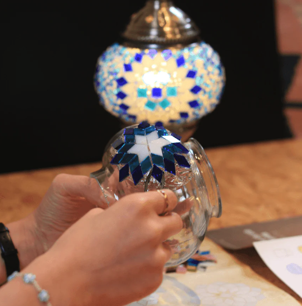 Person working on glass craft at Mosaic Art Studio with a blue and white mosaic design, with a decorative mosaic lamp in the background.