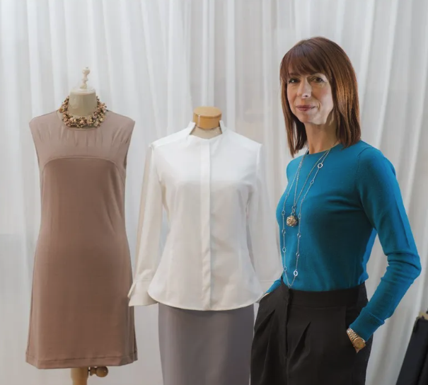 Kathleen Todoruk standing next to two mannequins dressed in clothing, one with a beige dress and necklace, the other with a white blouse and gray skirt, against a white curtain background.