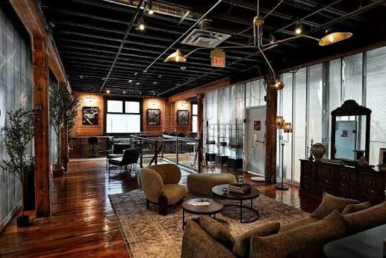 A spacious loft-style room with wooden floors, exposed brick walls, and large windows. The room is decorated with vintage furniture, including a sofa, armchairs, and coffee tables, with a bar or buffet cabinet and various decorative items. There are some plants and wall art, with a ping pong table visible in the background and pendant lights hanging from the ceiling.