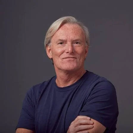 Older man with gray hair wearing a dark blue shirt, crossing his arms, standing against a gray background.