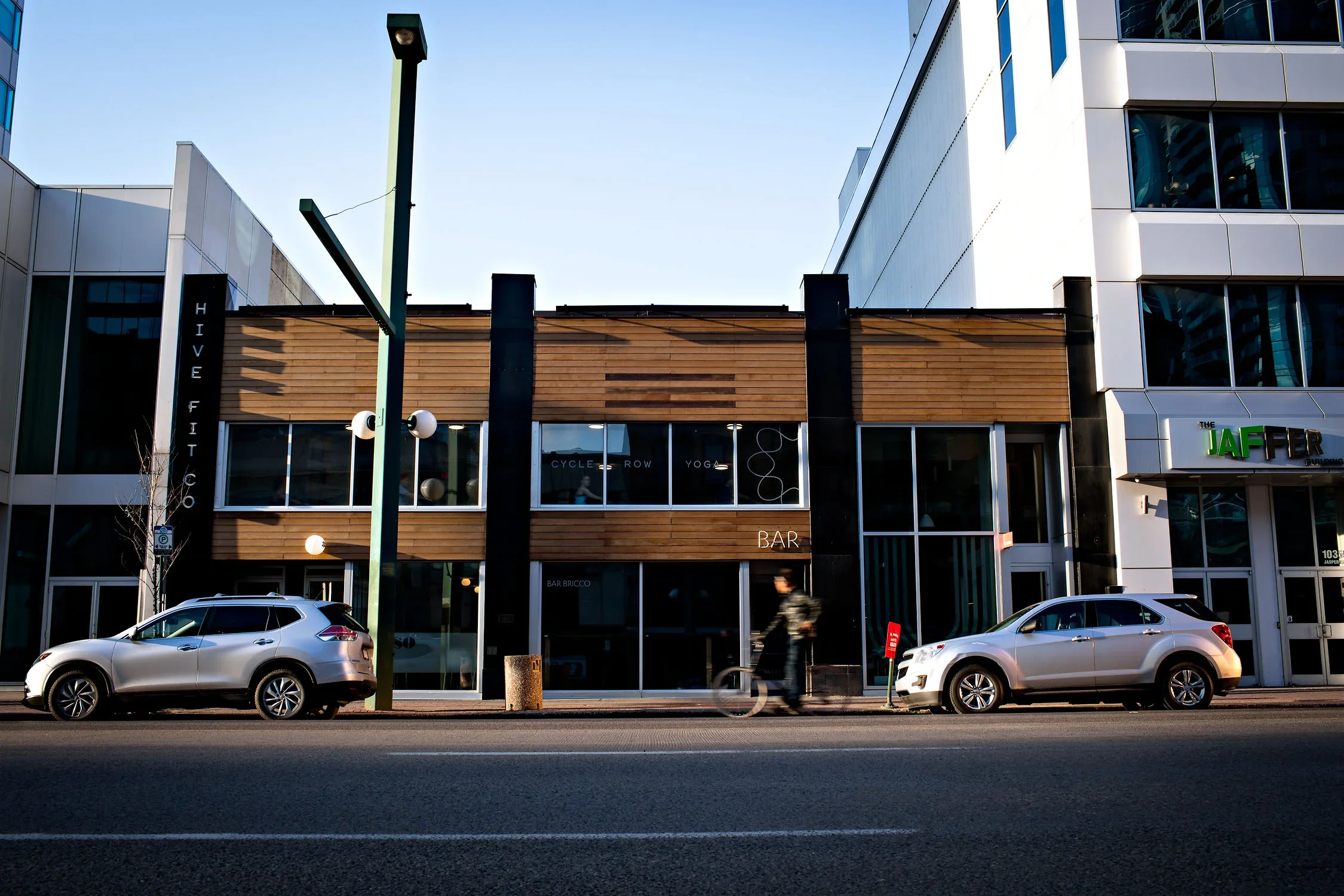 A city street scene with modern office buildings, cars parked along the curb, and a person walking past the buildings. The buildings feature a mix of glass and wood exterior finishes, and there are street lights and a parking sign visible.