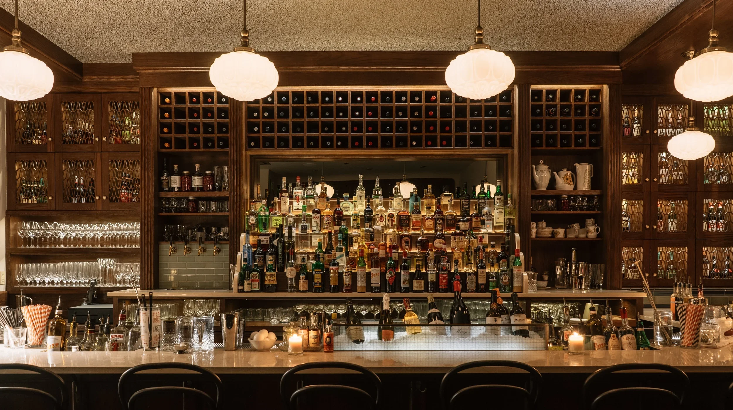 Clementine Bar with a wooden back wall filled with wine bottles, liquor bottles, glasses, and mugs. There are hanging pendant lights illuminating the bar area.