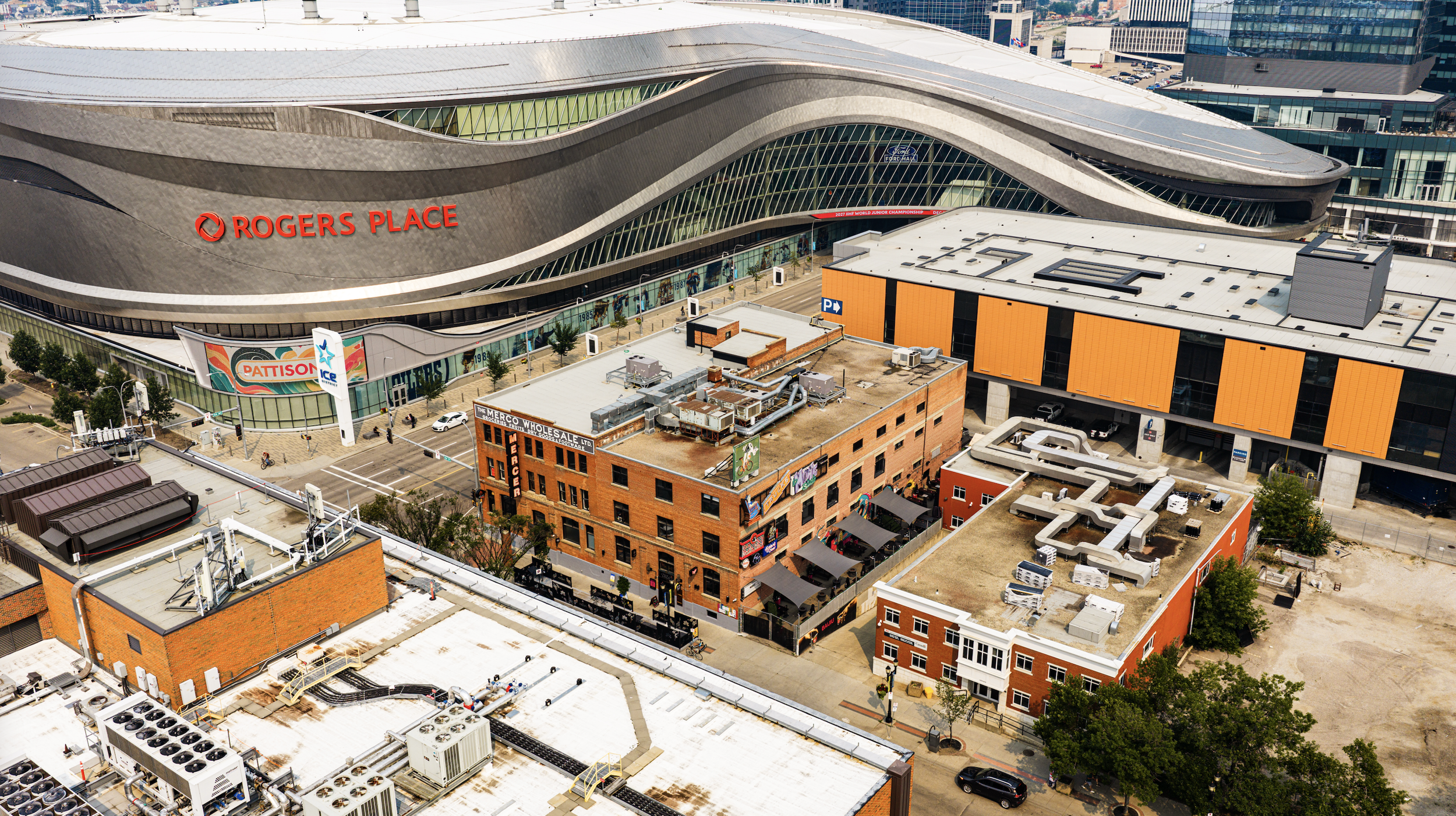 Aerial view of Rogers Place, an arena in Edmonton, Alberta, Canada, with surrounding city buildings, parking areas, and some construction or maintenance equipment on the rooftops and nearby streets.