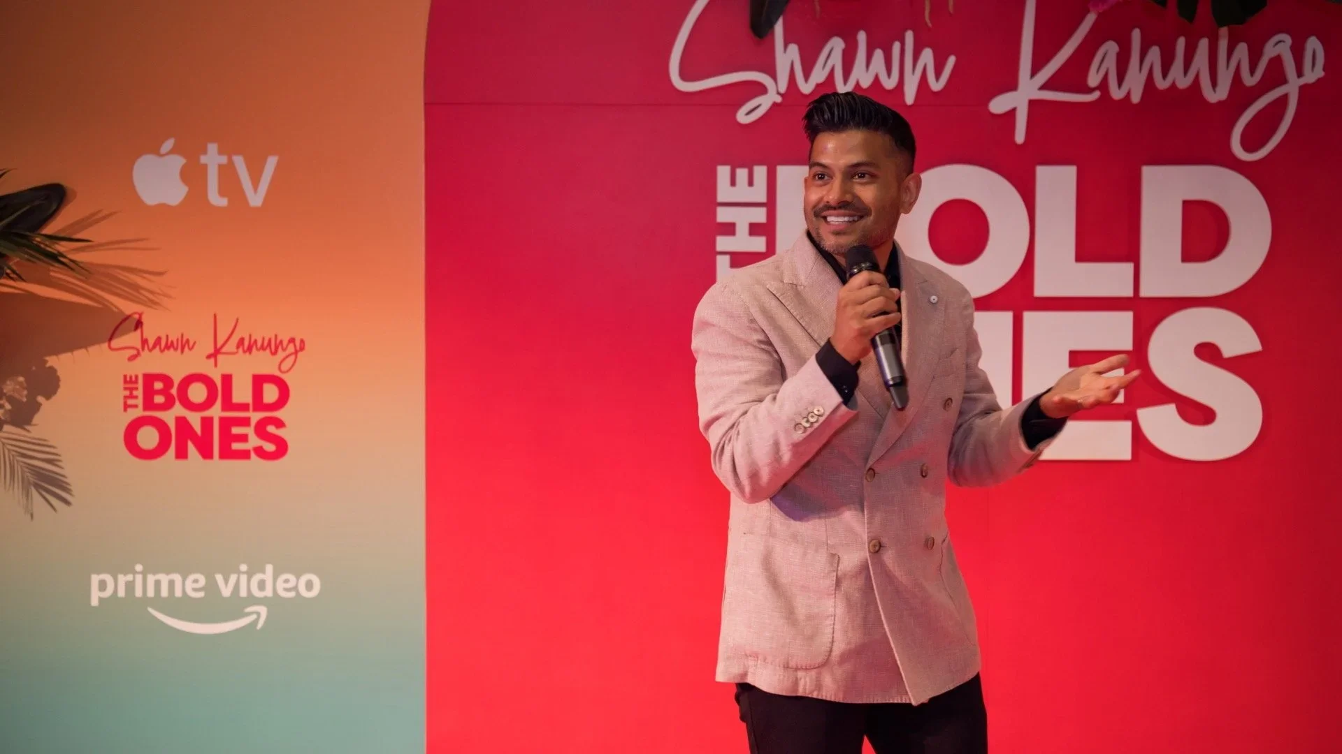 Shawn Kanugo in tan blazer holding microphone, smiling, speaking at a presentation with red background and logos for Apple TV, Prime Video, and The Bold Type.