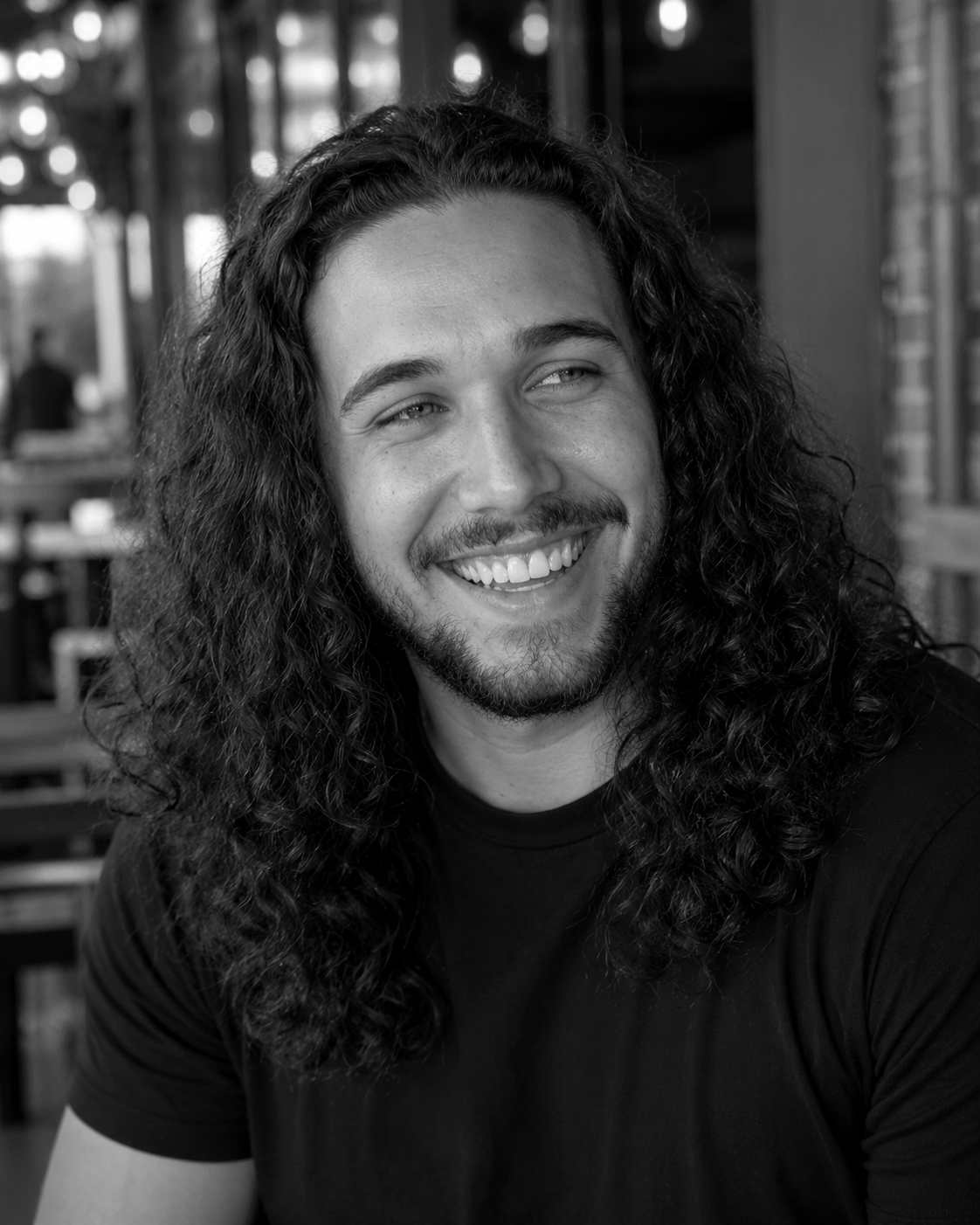 A smiling man with long curly hair and a beard, wearing a dark t-shirt, sitting in an outdoor cafe or patio area, captured in black and white.
