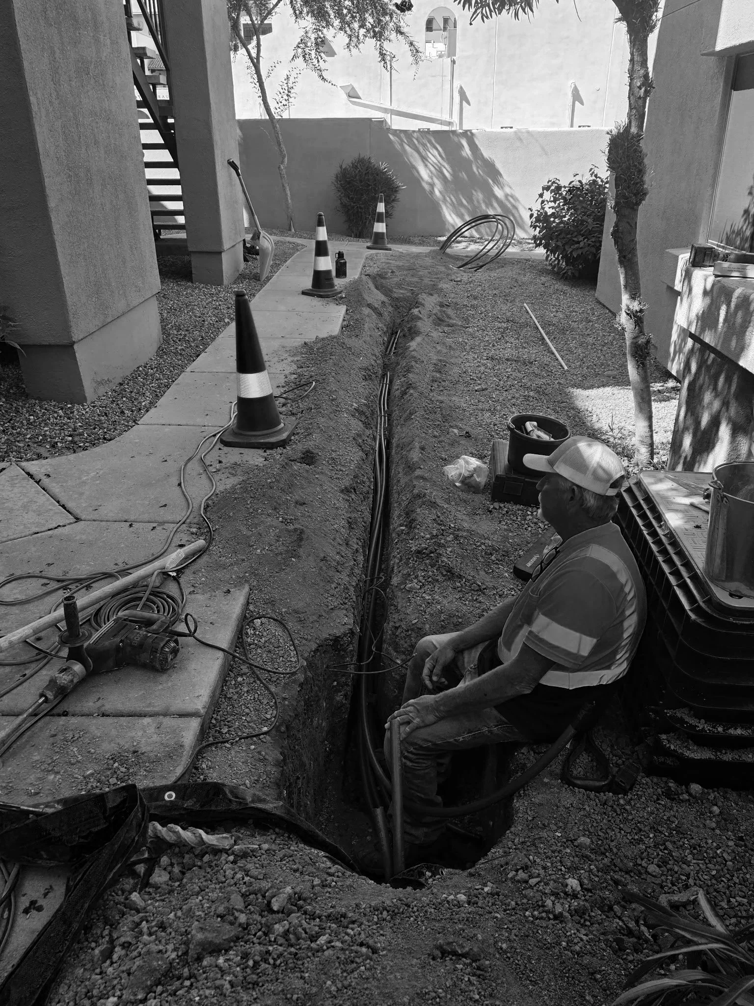 Construction worker sitting beside a trench with electrical wiring, construction cones, and tools in an outdoor area under sunlight.