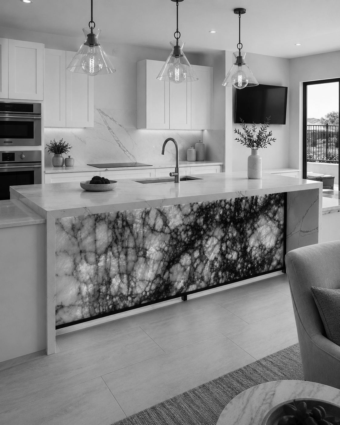 Modern kitchen with white cabinets, marble countertops, and a marble island with a black and white veined pattern. Hanging pendant lights above the island, a vase with greenery, and a bowl of fruit.