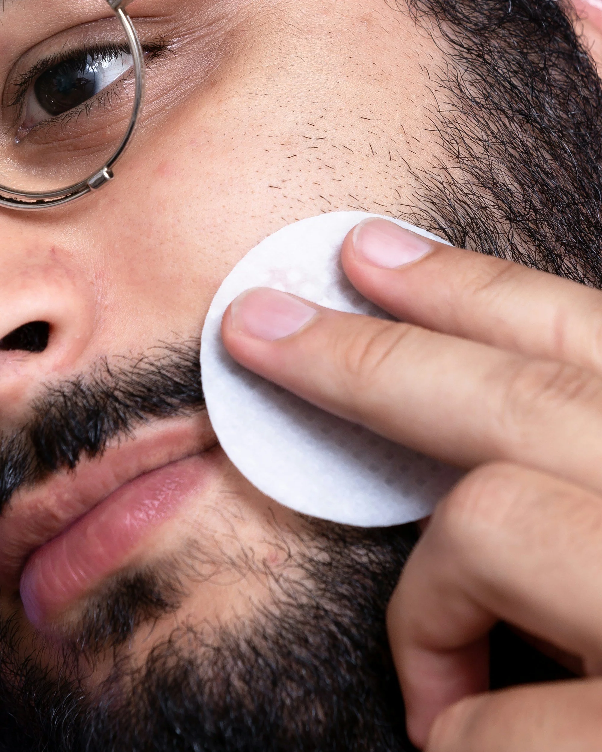 Man with glasses wiping his cheeks with a cotton pad above his beard