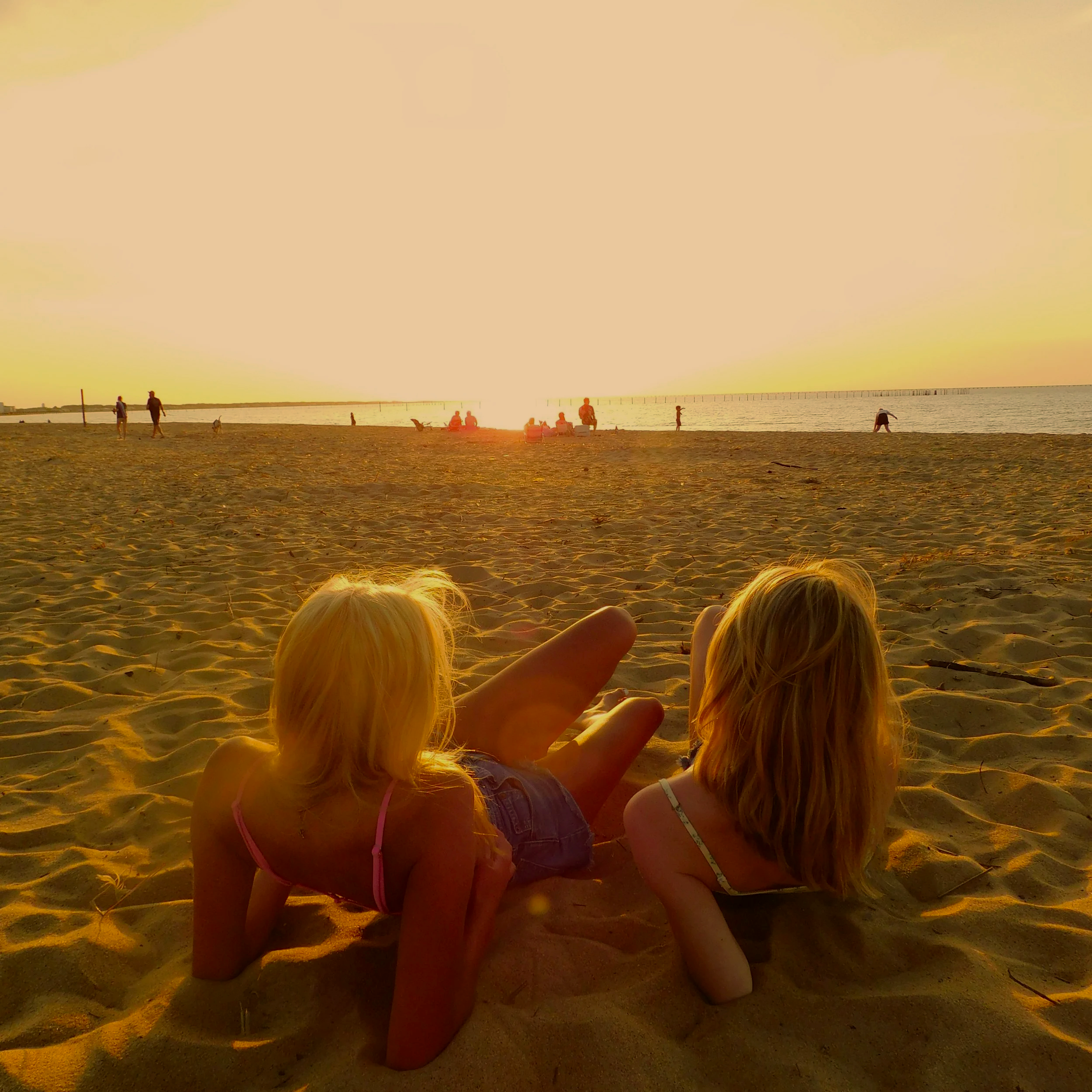Two children with blonde hair sitting on the sand at sunset, facing the water, with other people in the background enjoying the beach.