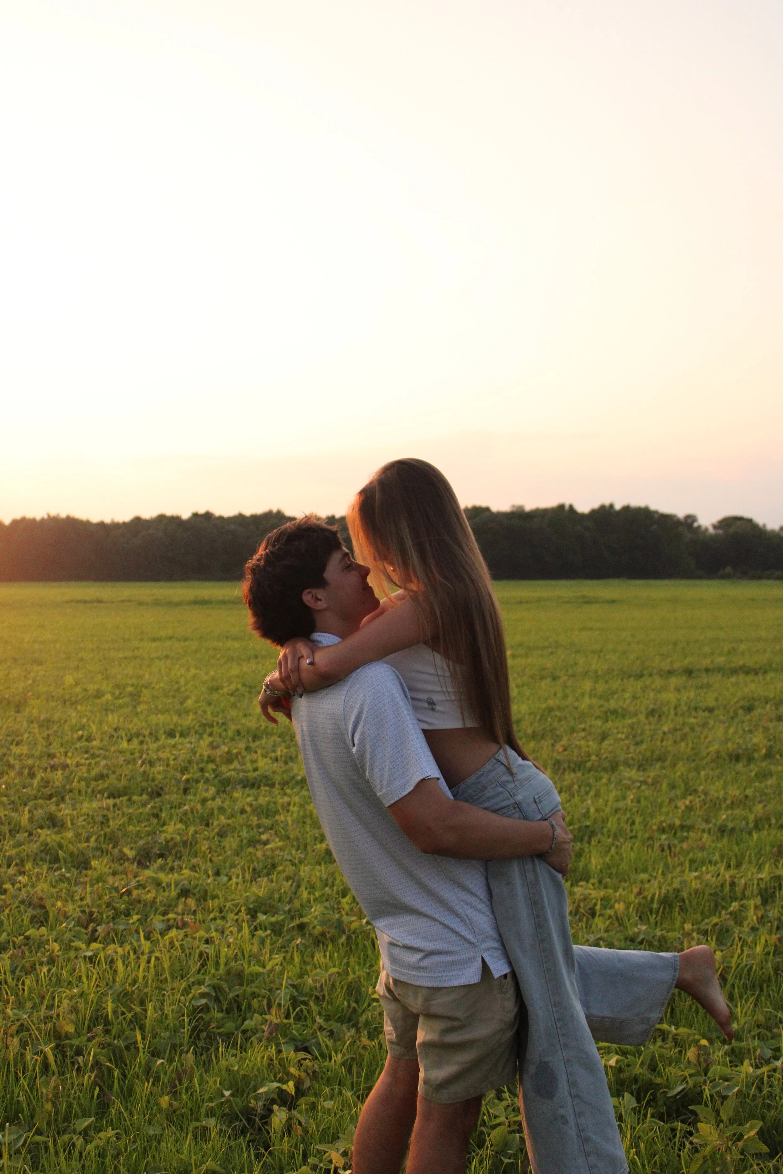 A young man lifting a young woman in a field at sunset, as they embrace and look at each other affectionately.