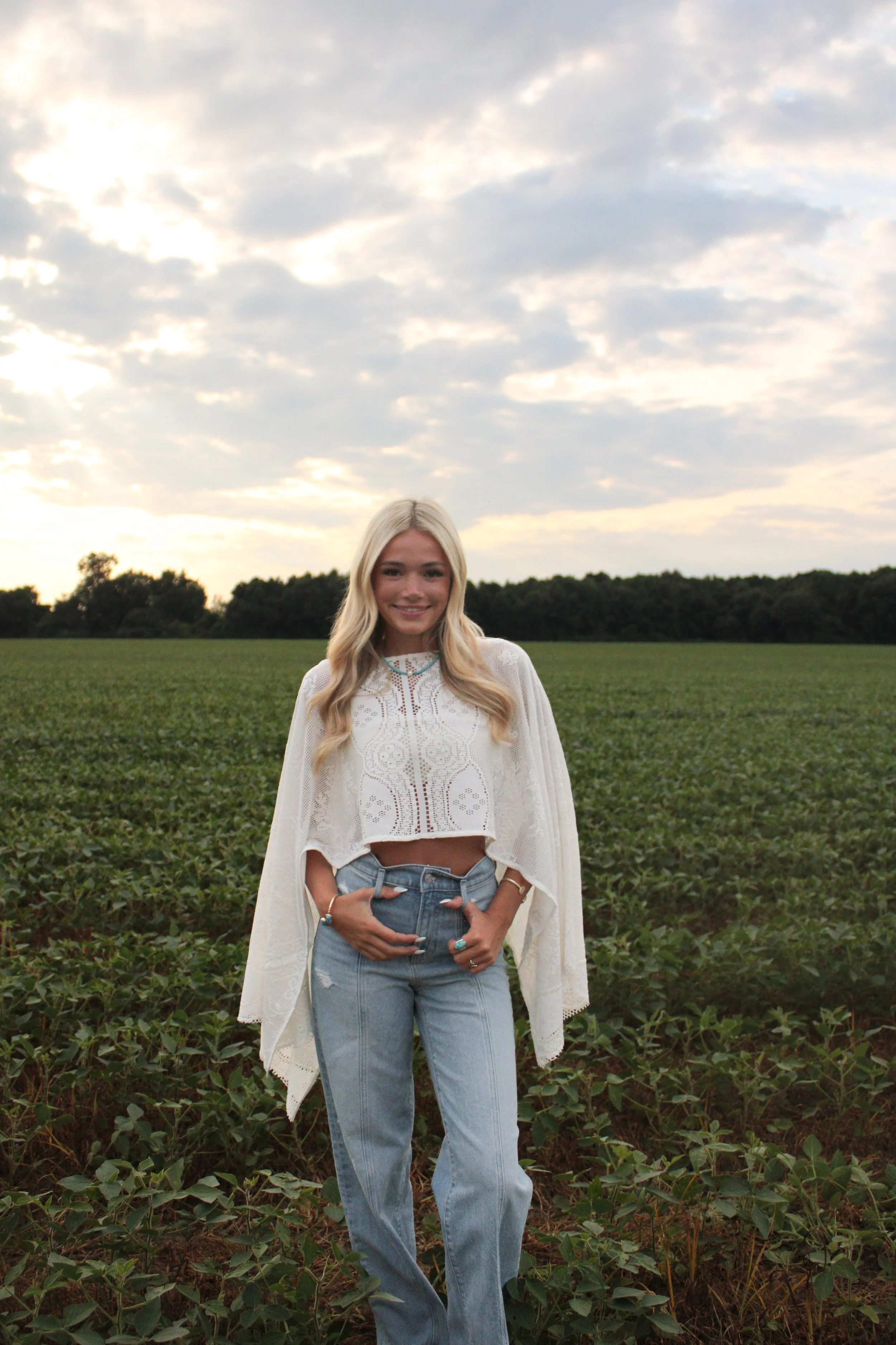 A young woman with long blonde hair smiling, standing in a green field with trees in the distance during cloudy weather.