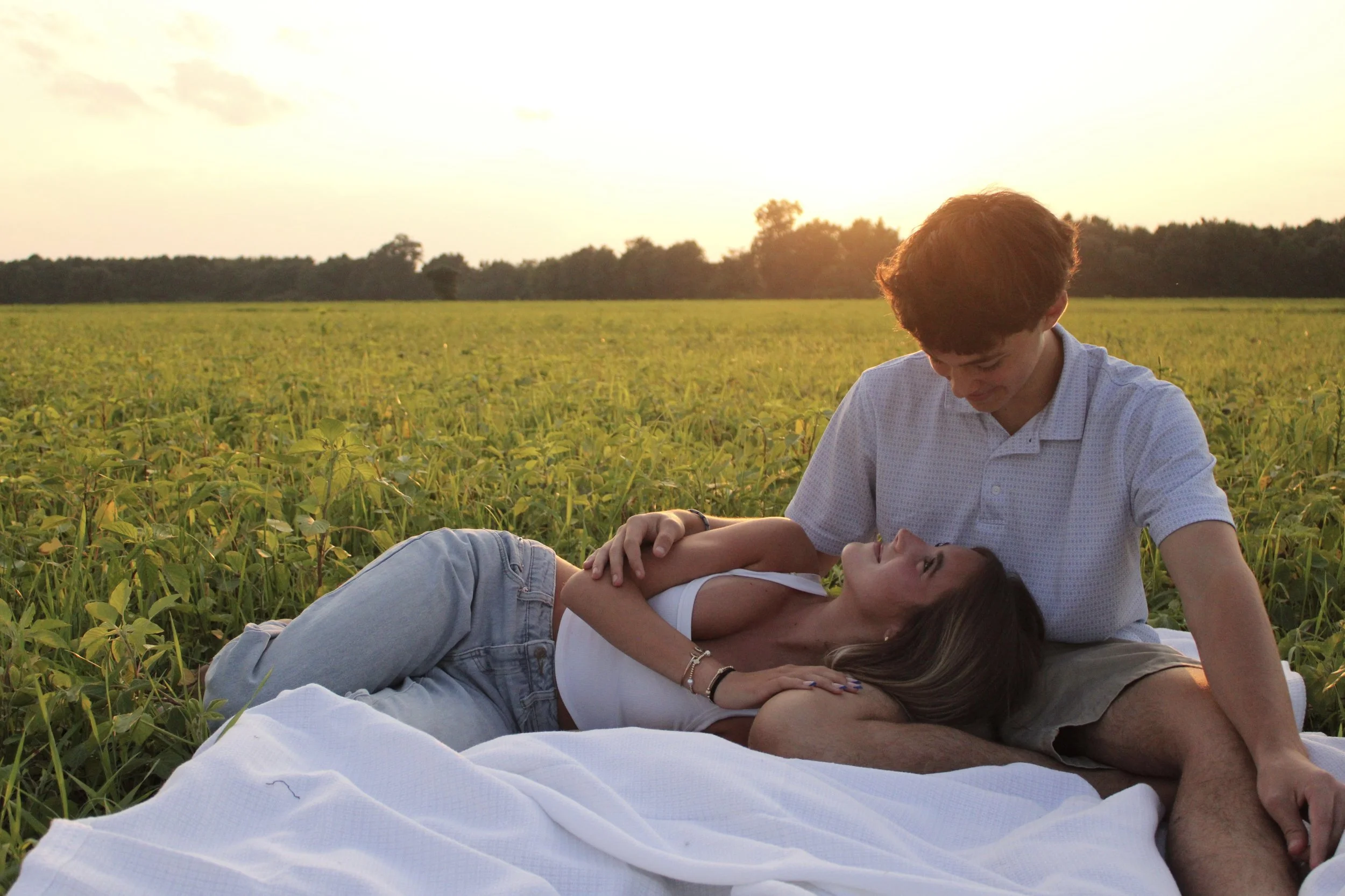 Two young adults, a man and a woman, lying and sitting in a green field during sunset. The man is gently touching the woman's face as she rests with her eyes closed. They are surrounded by lush plants, with trees and the sky in the background.
