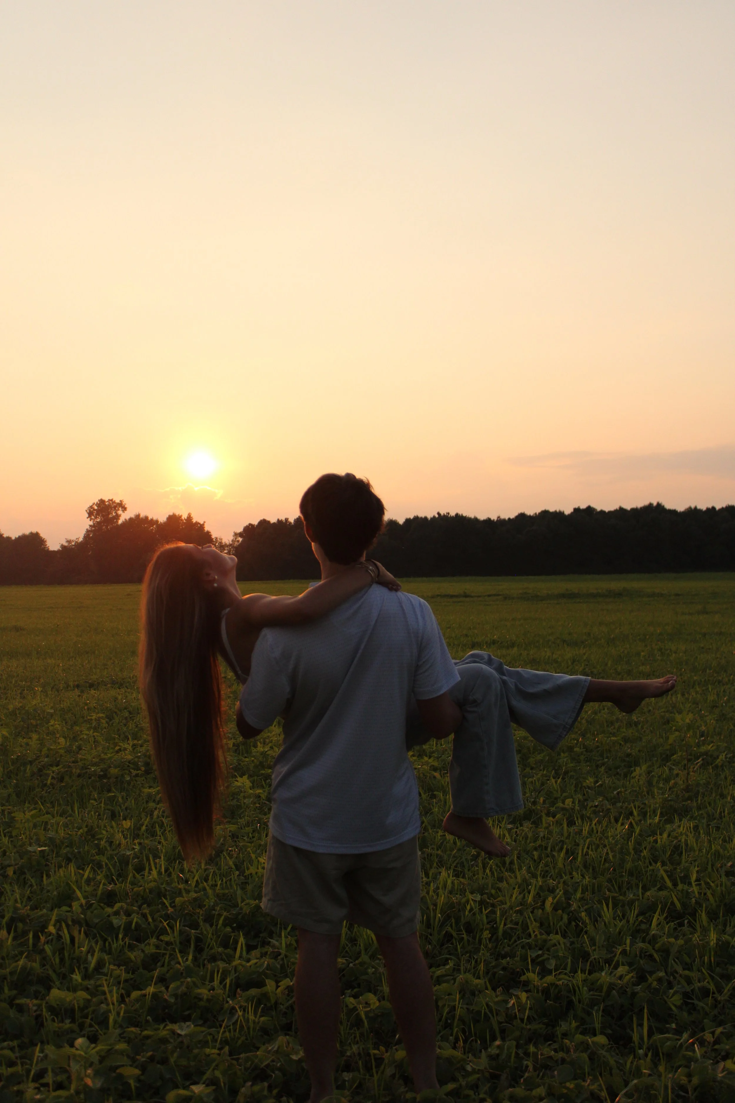 A couple sitting in a grassy field at sunset, with the woman embracing the man and the woman being carried over the man's shoulder, surrounded by trees and a clear sky.
