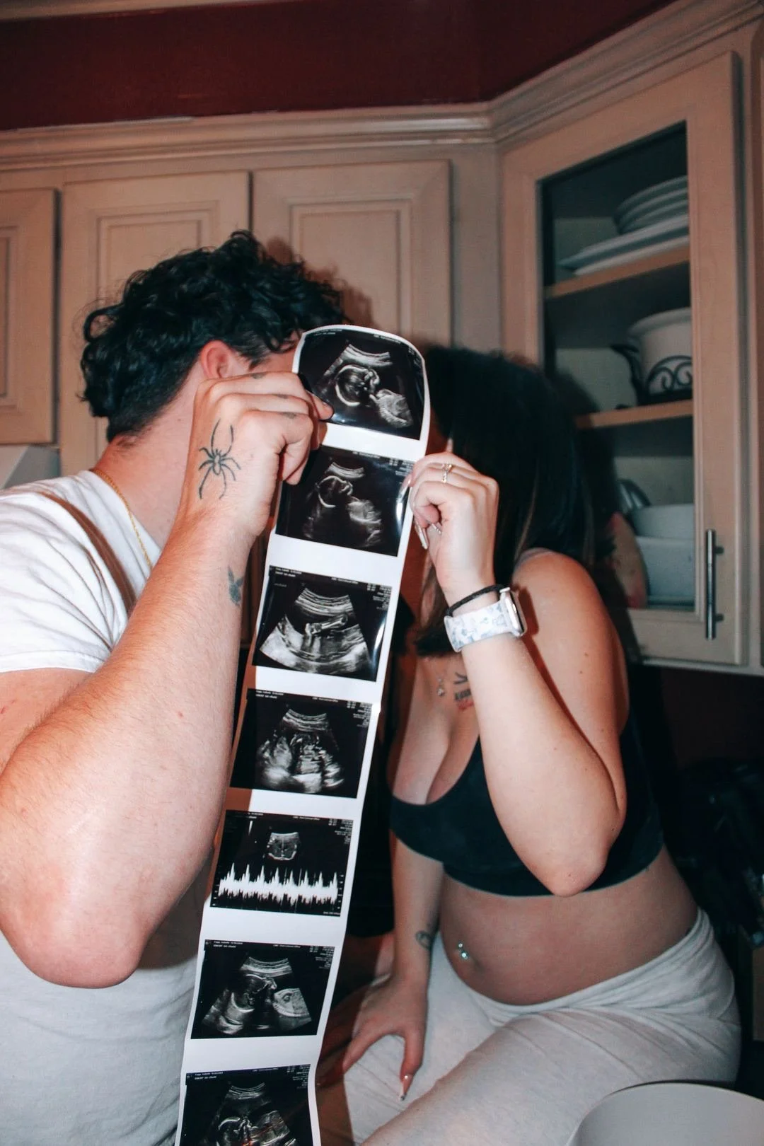 A man and woman are in a kitchen looking at ultrasound images, with the man holding the ultrasound photos and the woman wearing a tank top and sitting on a counter.