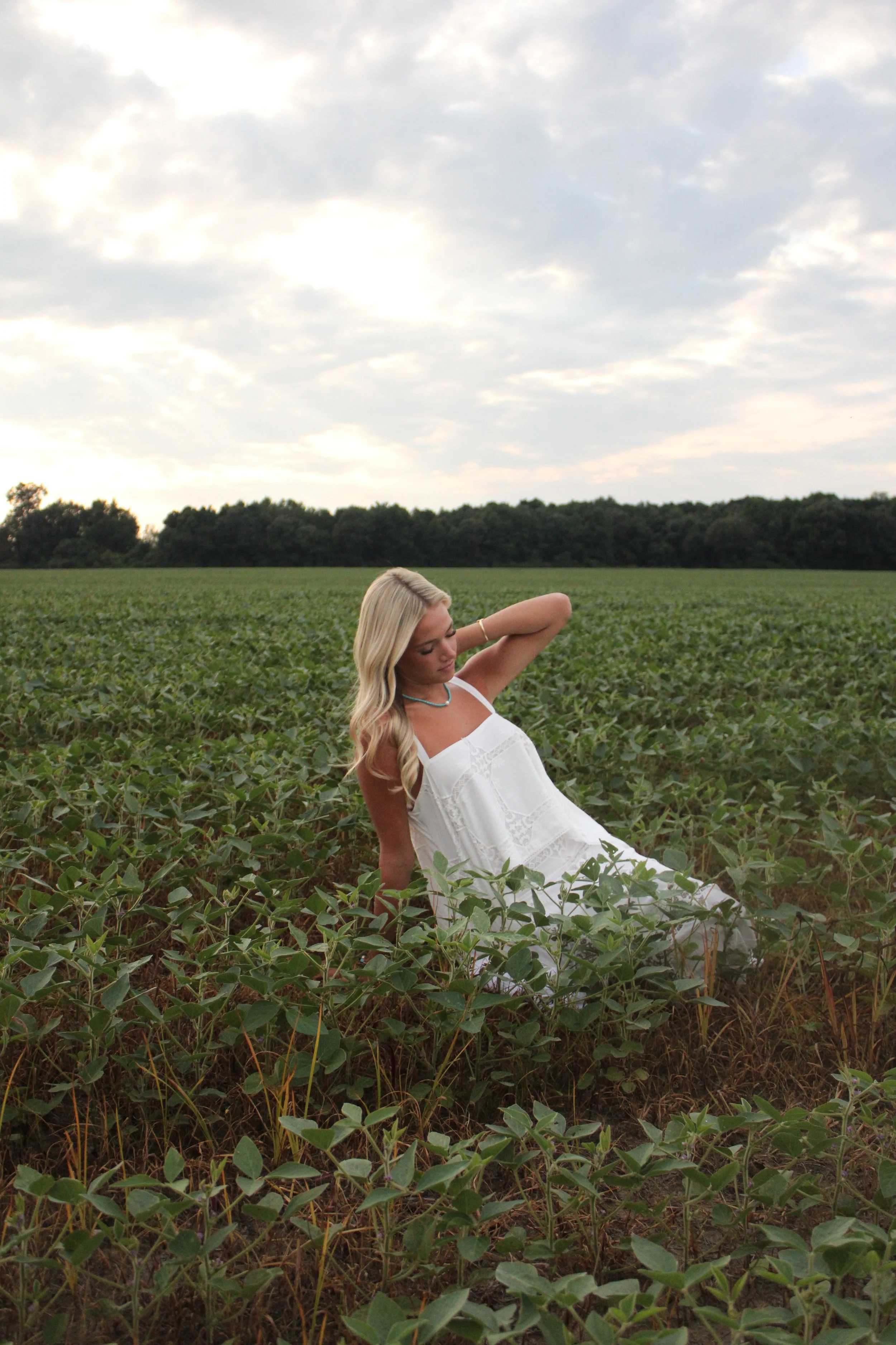A woman in a white dress sitting in a lush green field with a line of trees in the background and a cloudy sky overhead.