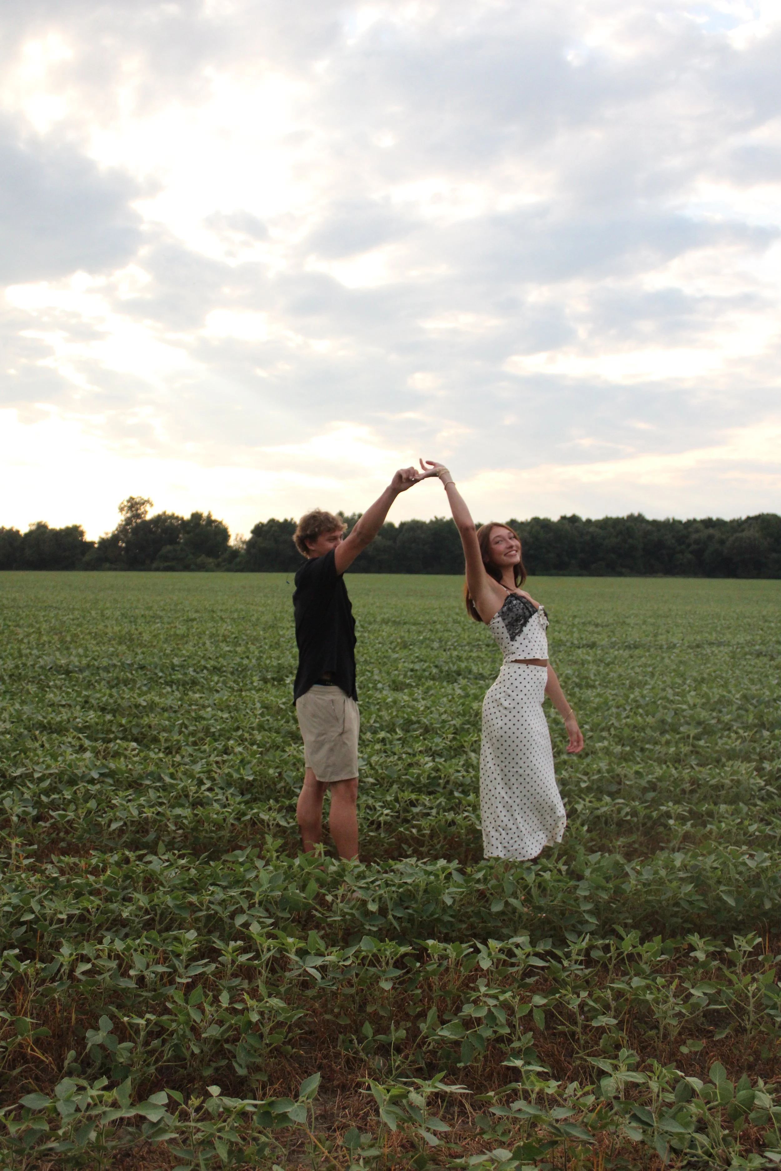 A young man and woman are standing in a green field under a cloudy sky. They are dancing, holding hands above their heads, and smiling at each other.