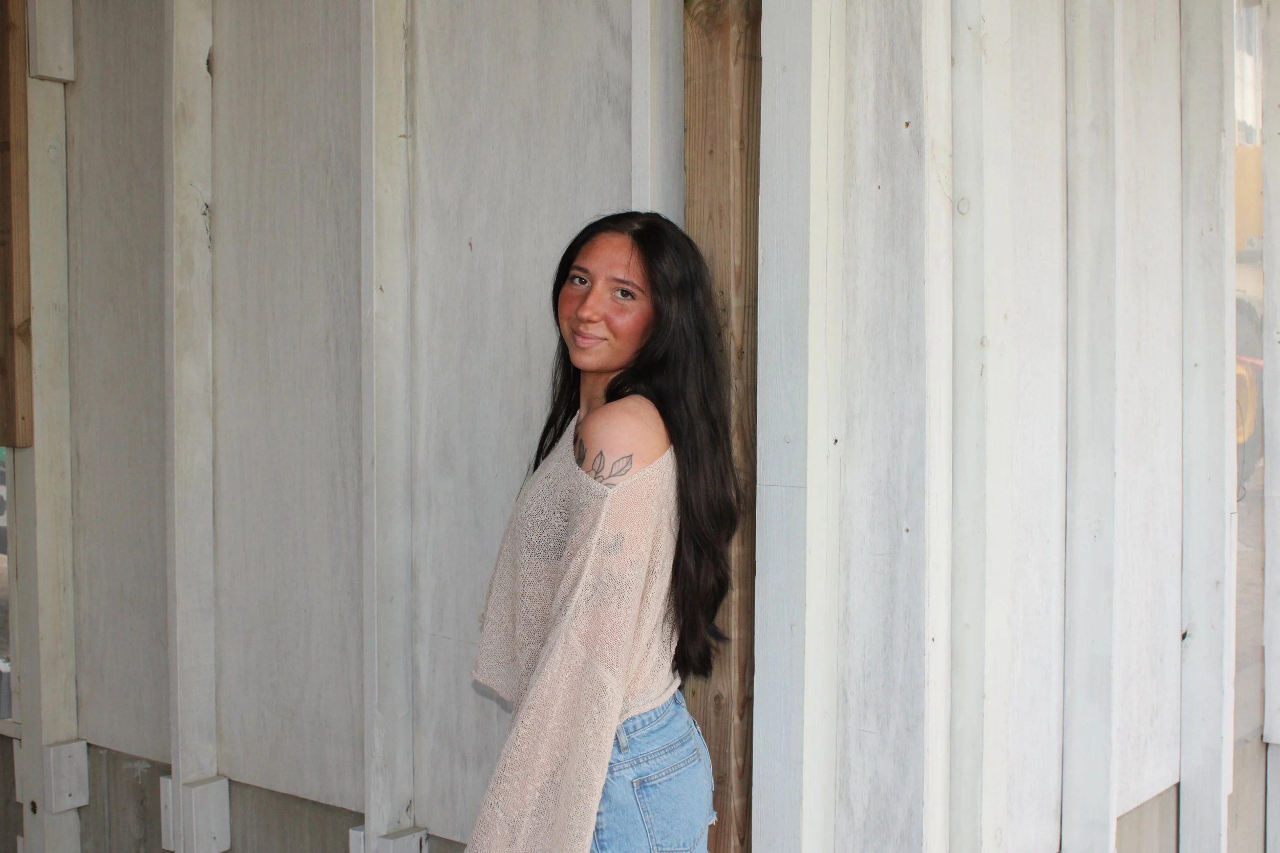 A woman with long dark hair, light brown skin, and a flower tattoo on her shoulder, standing against a white wooden fence with vertical planks and some exposed wooden beams.