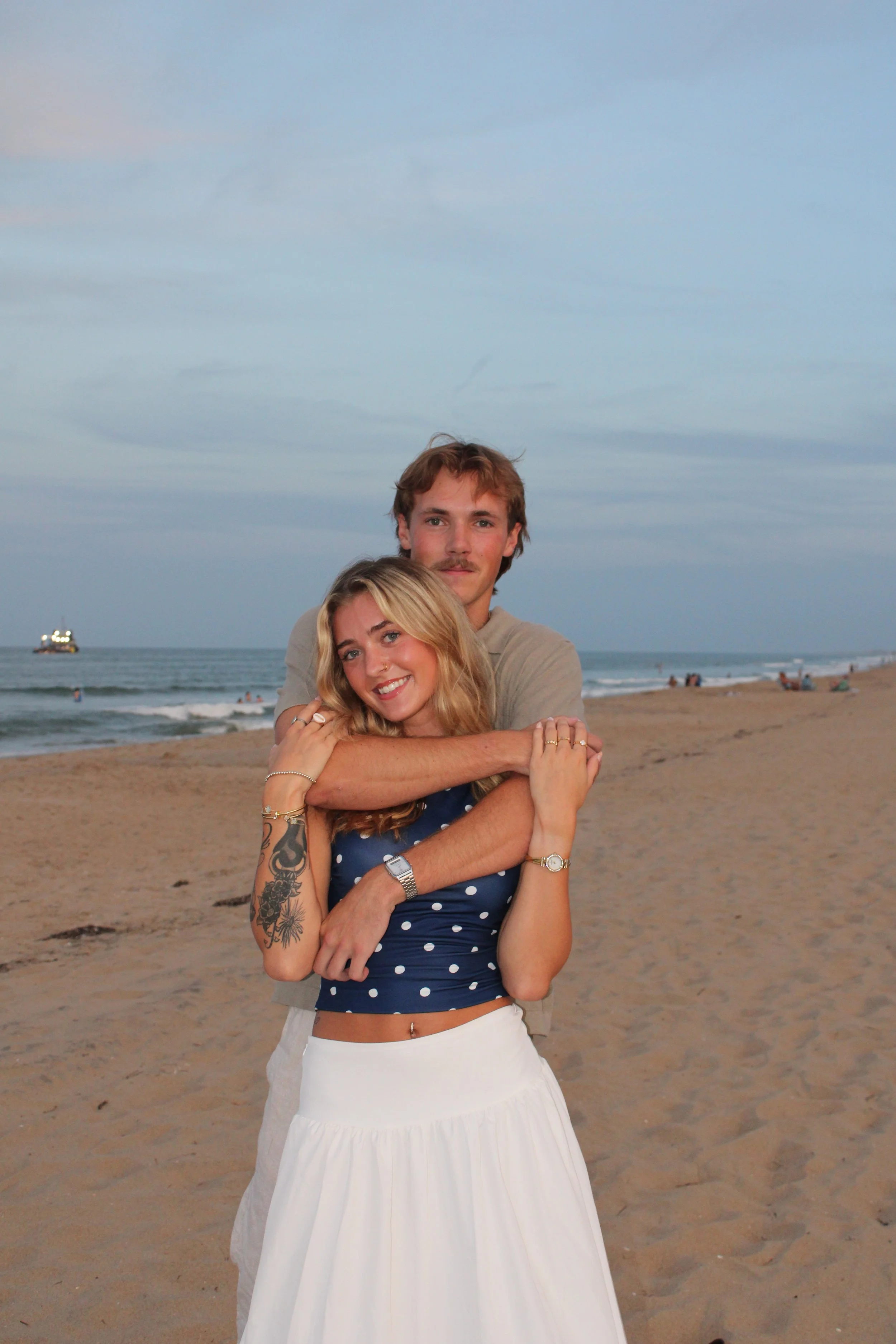 A smiling couple stands closely together on a beach, with the ocean and a boat on the water in the background, during dusk.