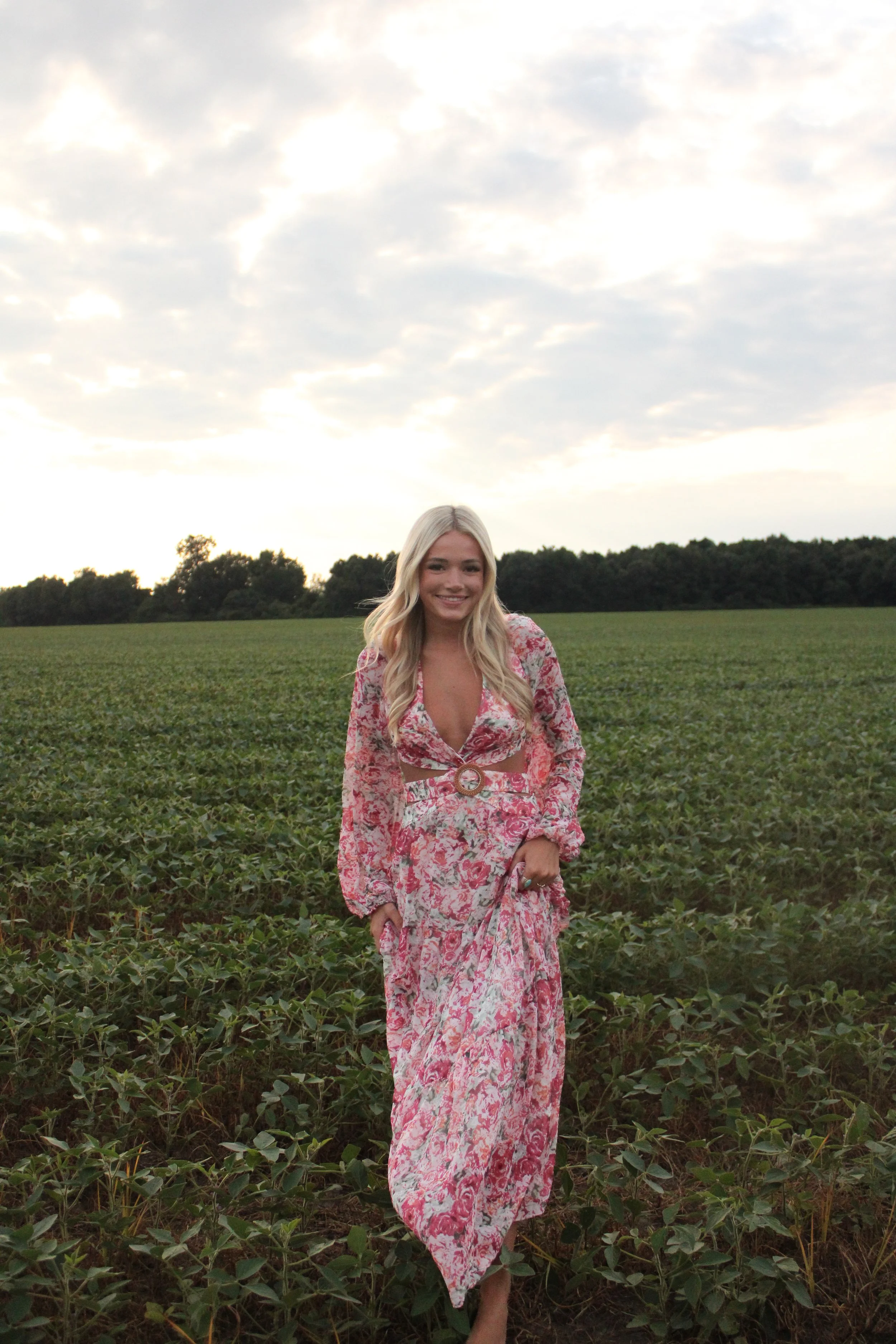 A young woman with long blond hair smiling and wearing a pink floral dress, standing in a green field with trees in the background and a cloudy sky overhead.