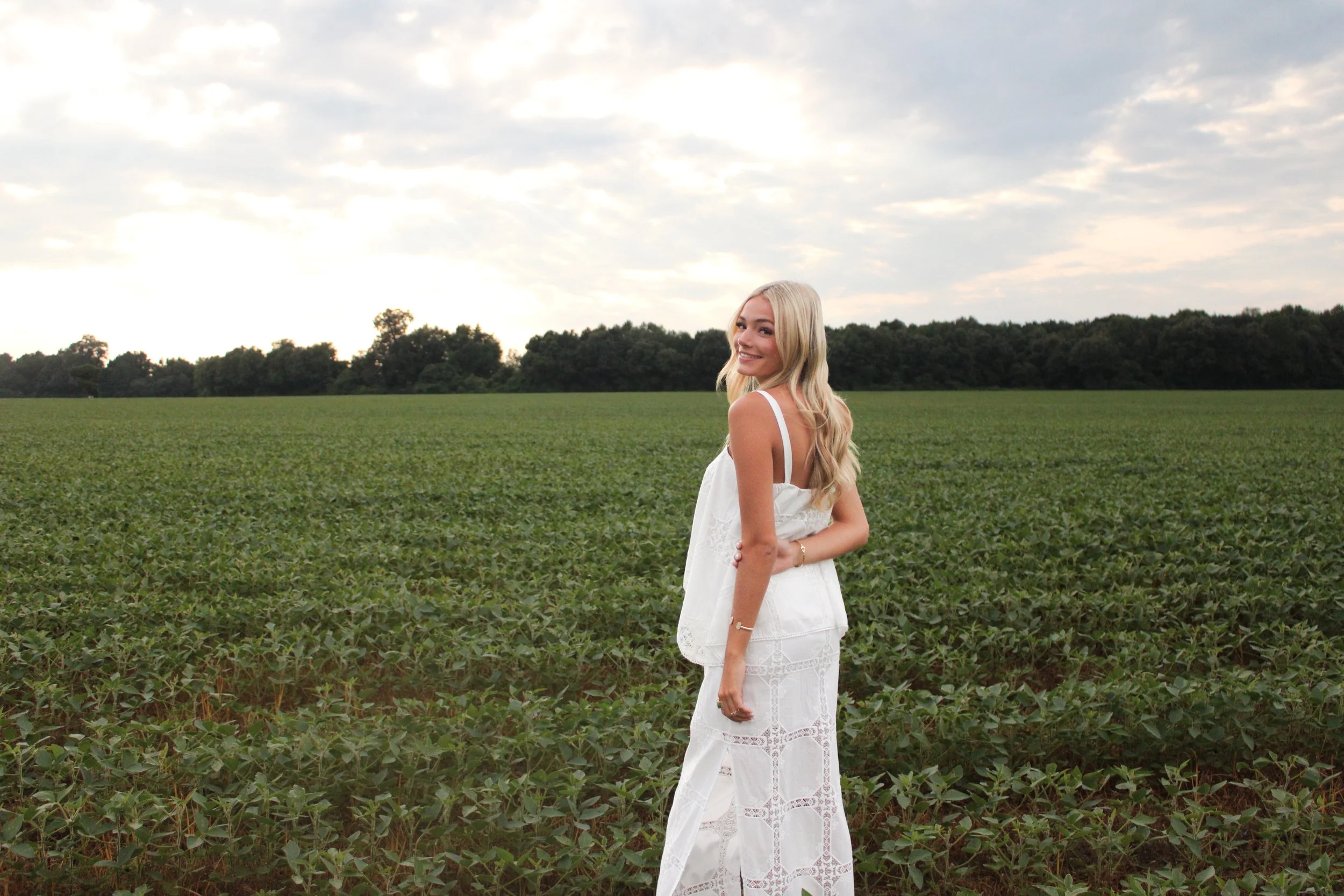 A young woman with long blonde hair wearing a white dress with intricate lace details, standing in a green field under a partly cloudy sky, smiling and looking back at the camera.