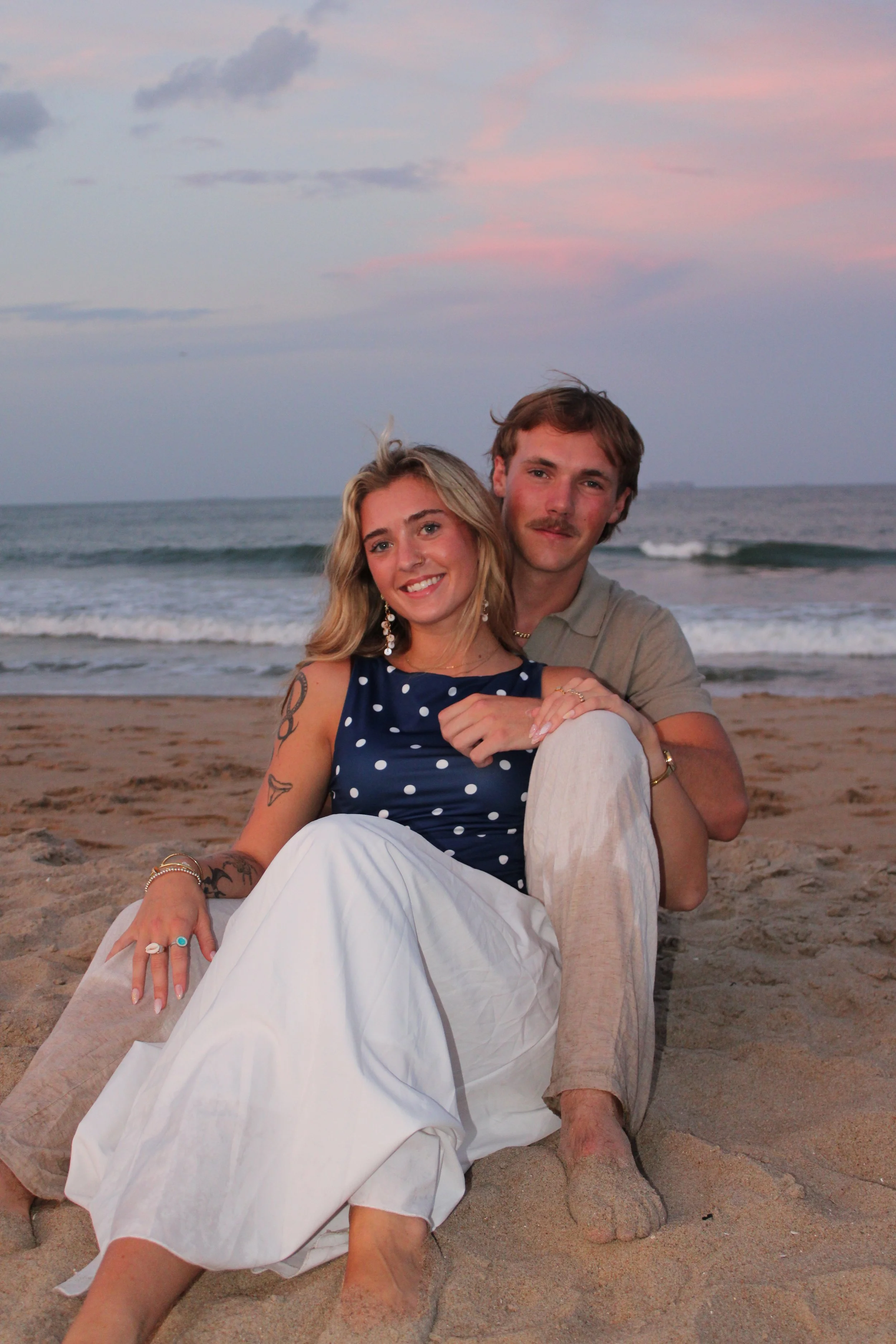 A young woman and a young man sitting on the sandy beach at sunset, smiling at the camera. The woman has blonde hair, tattoos on her arms, and is wearing a navy polka-dot top. The man has light brown hair, a mustache, and is wearing a beige shirt. Th
