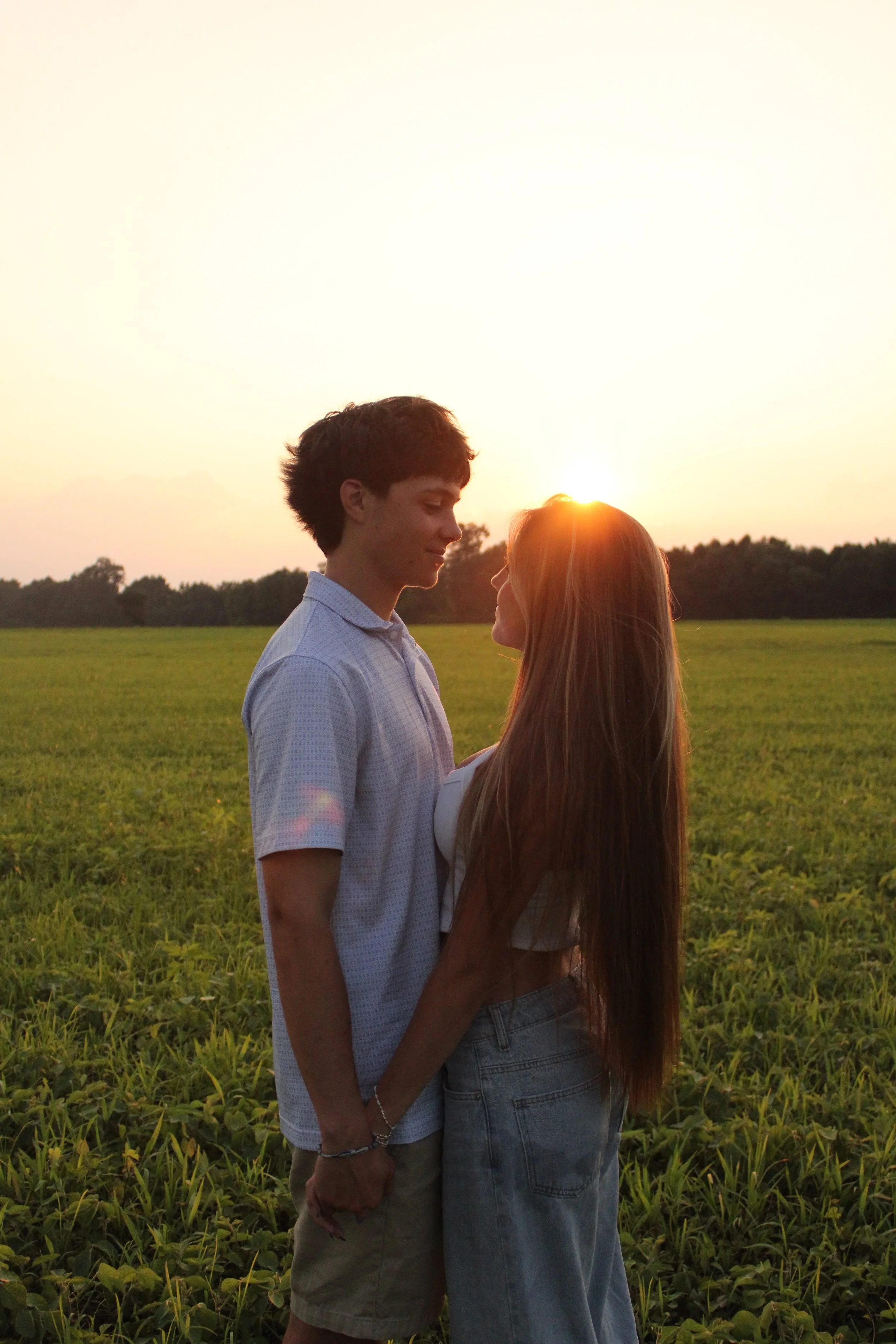 A young man and woman are standing outdoors in a field at sunset, facing each other and holding hands, with the sun setting behind them.