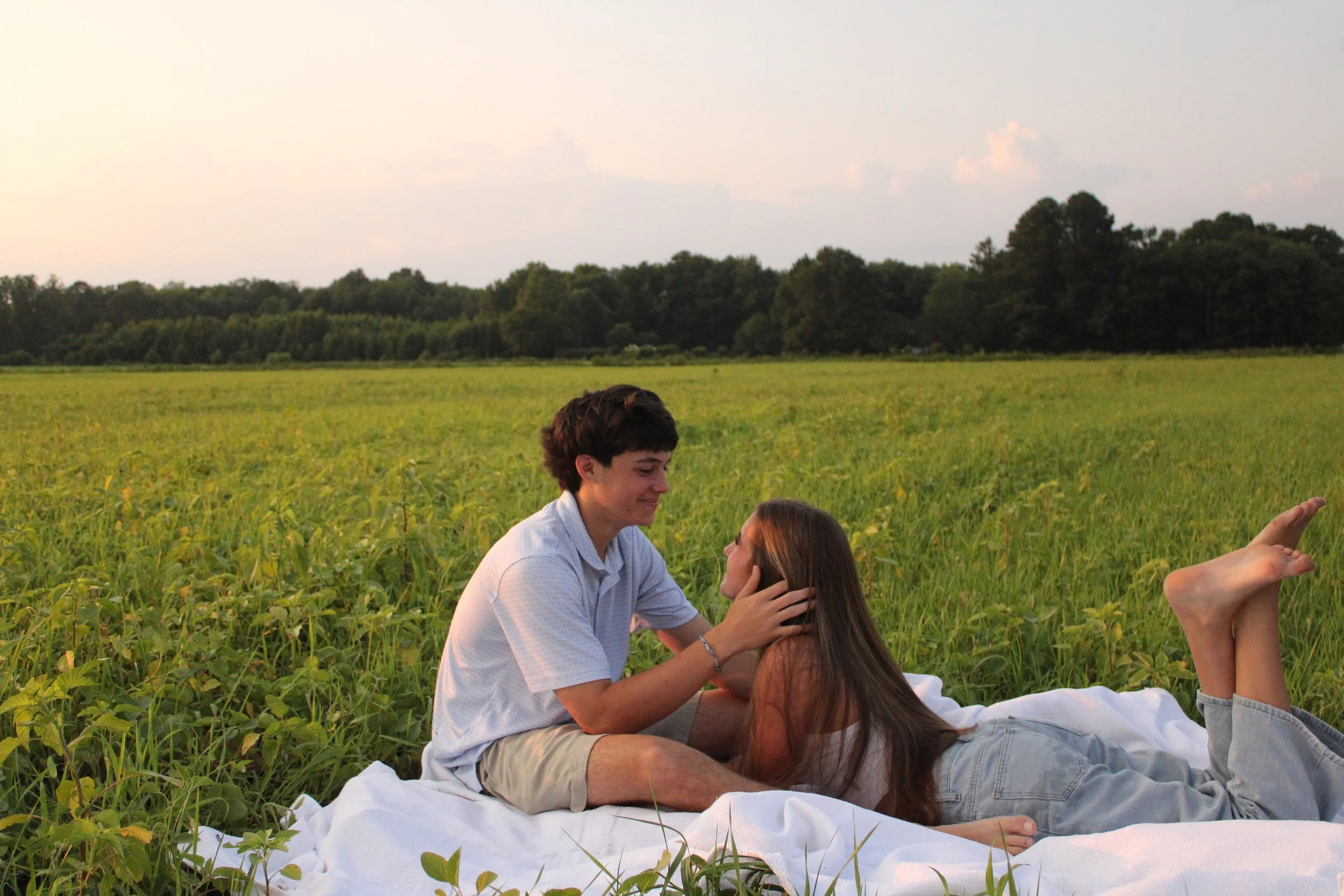 A young couple sitting on a white blanket in a green field, with the man gently holding the woman's face as they look at each other, surrounded by trees in the background during sunset.