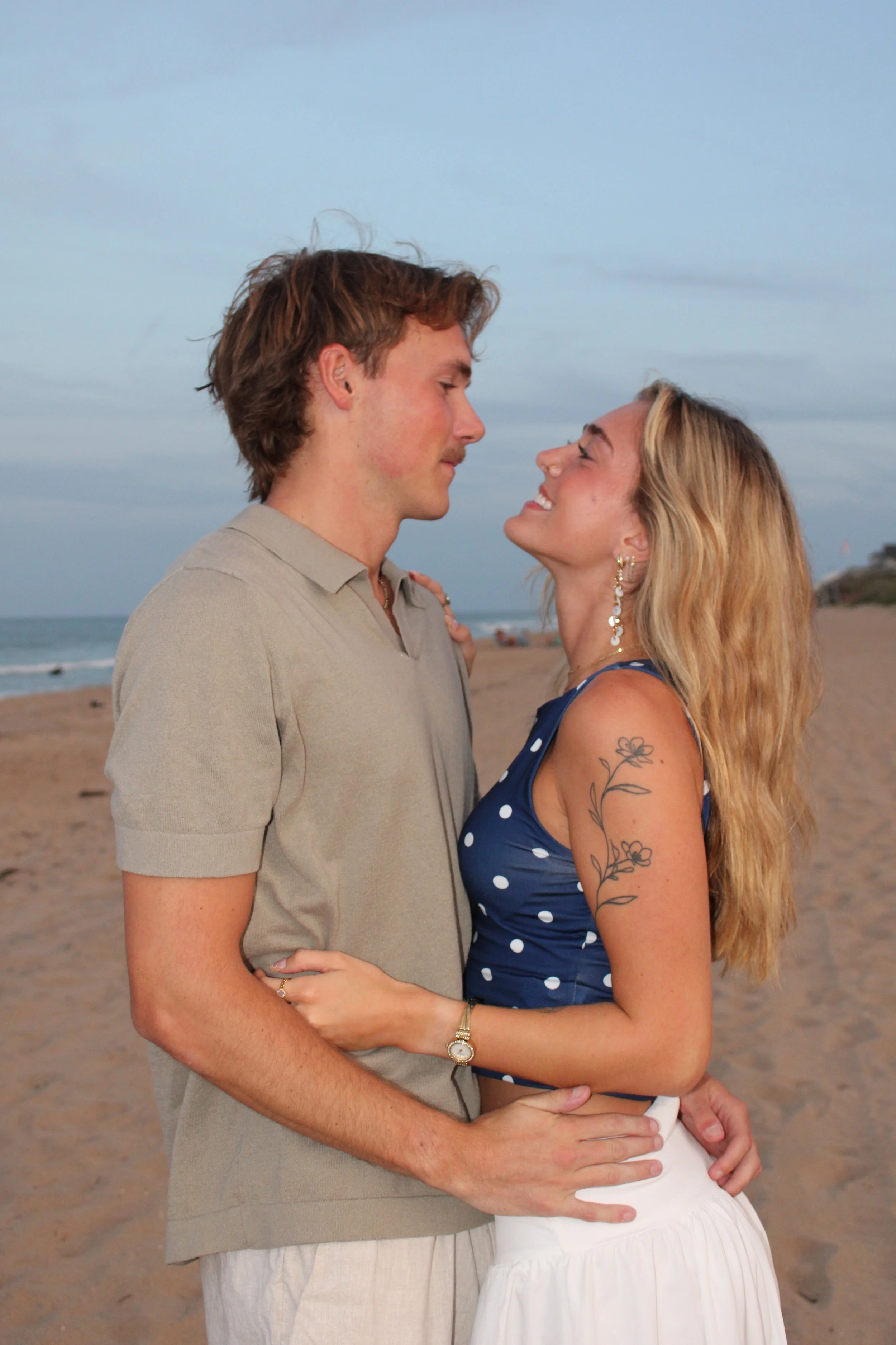 A couple is standing on a sandy beach, facing each other closely with smiles. The man has brown hair and is wearing a beige shirt with beige pants, while the woman has long blonde hair, tattoos on her arm, and is wearing a blue polka-dot top and a wh