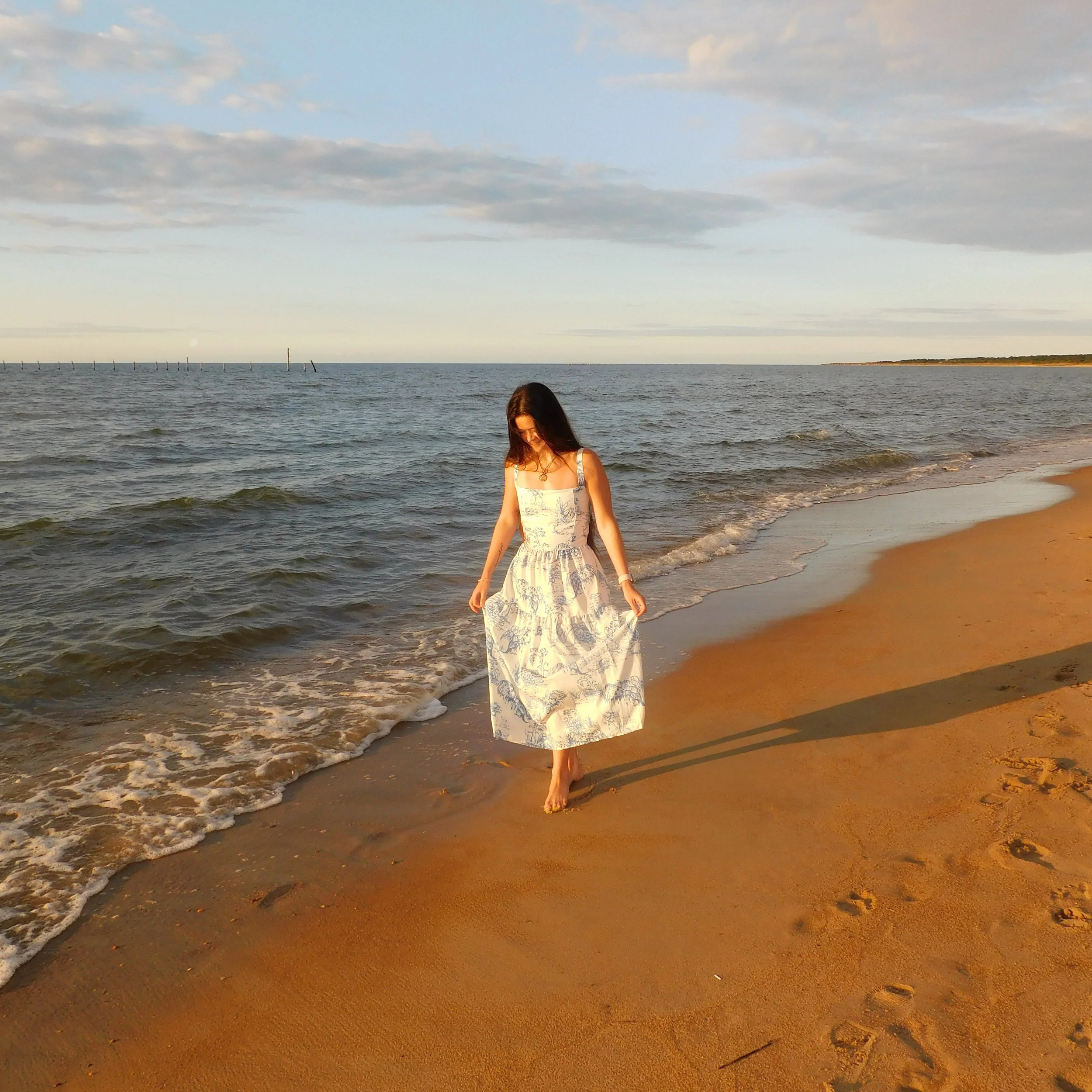 A woman in a white dress with blue patterns walking along the sandy beach near the water's edge during sunset or sunrise, with the ocean and partly cloudy sky in the background.