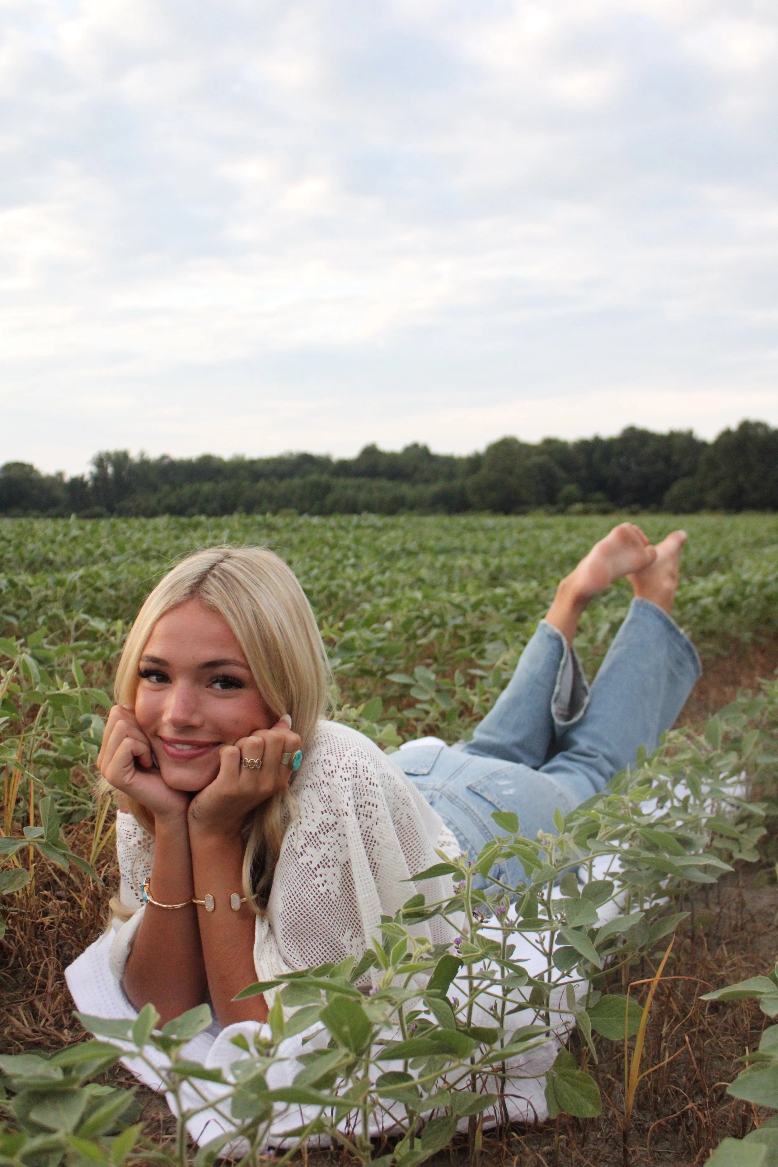 Young woman with blonde hair lying on her stomach in a soybean field, smiling and resting her chin on her hands, with another person laying upside down in the background, all outdoors under a cloudy sky.