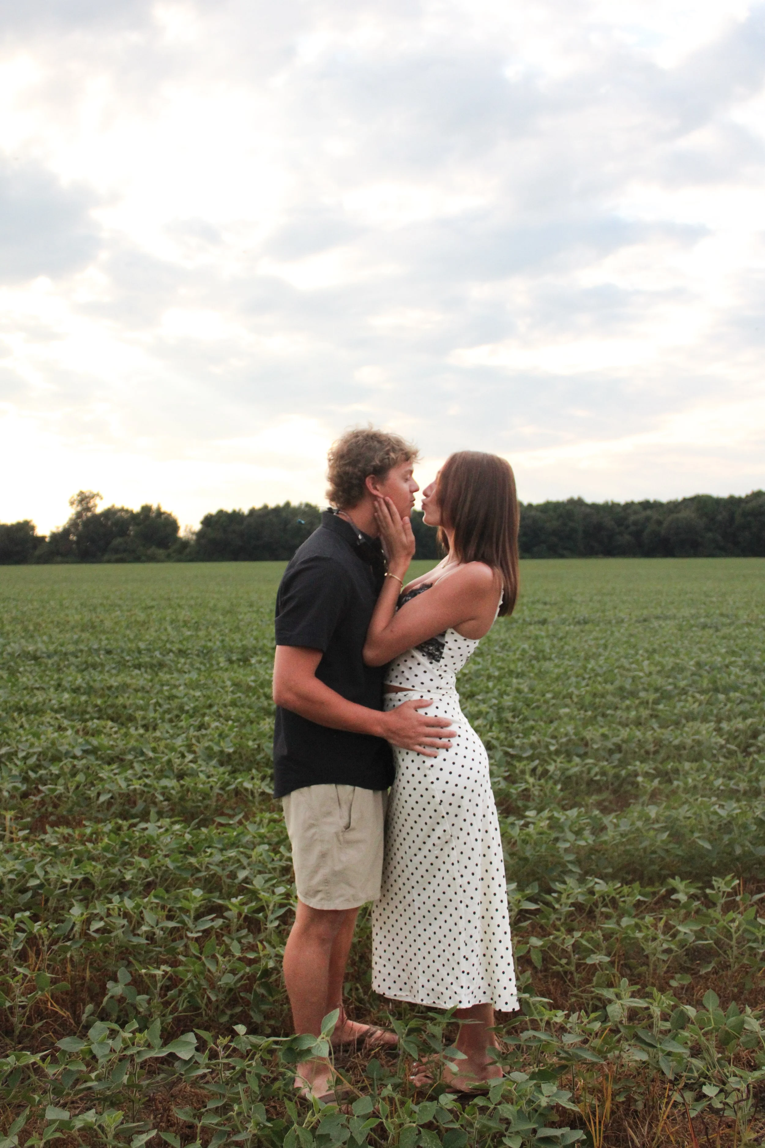 A young couple stands in a field of green plants, facing each other and touching faces, with trees and a partly cloudy sky in the background.